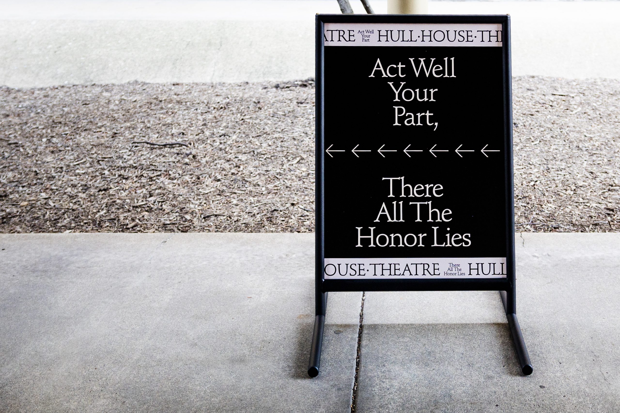 A black sandwich board sign with white text reads, “Act Well Your Part, There All The Honor Lies,” and is positioned on a sidewalk beside a dirt area. The sign is for Hull House Theatre.