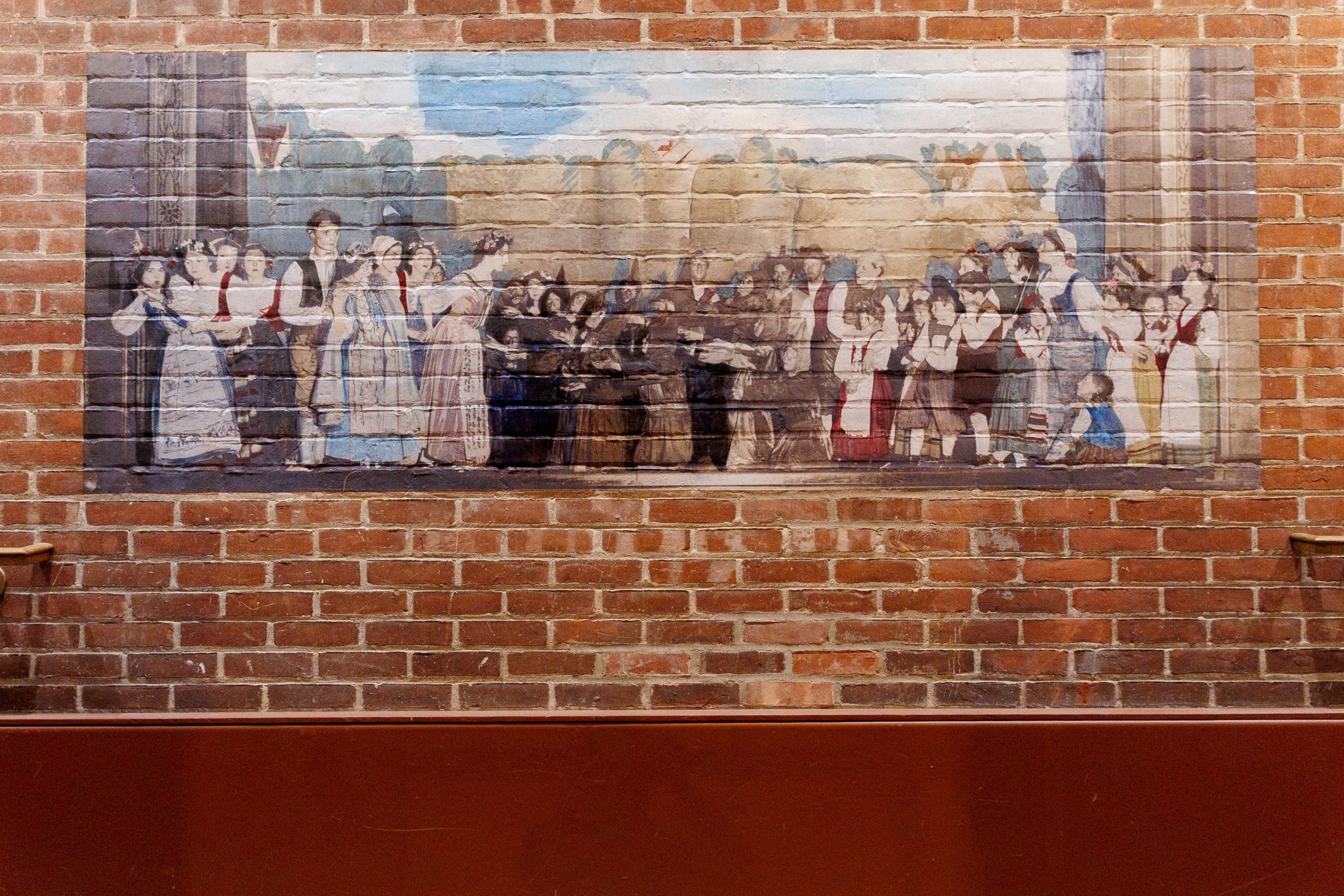 A mural painted on a brick wall depicts a group of people in historical clothing gathered and celebrating outside, with blue sky and trees in the background.