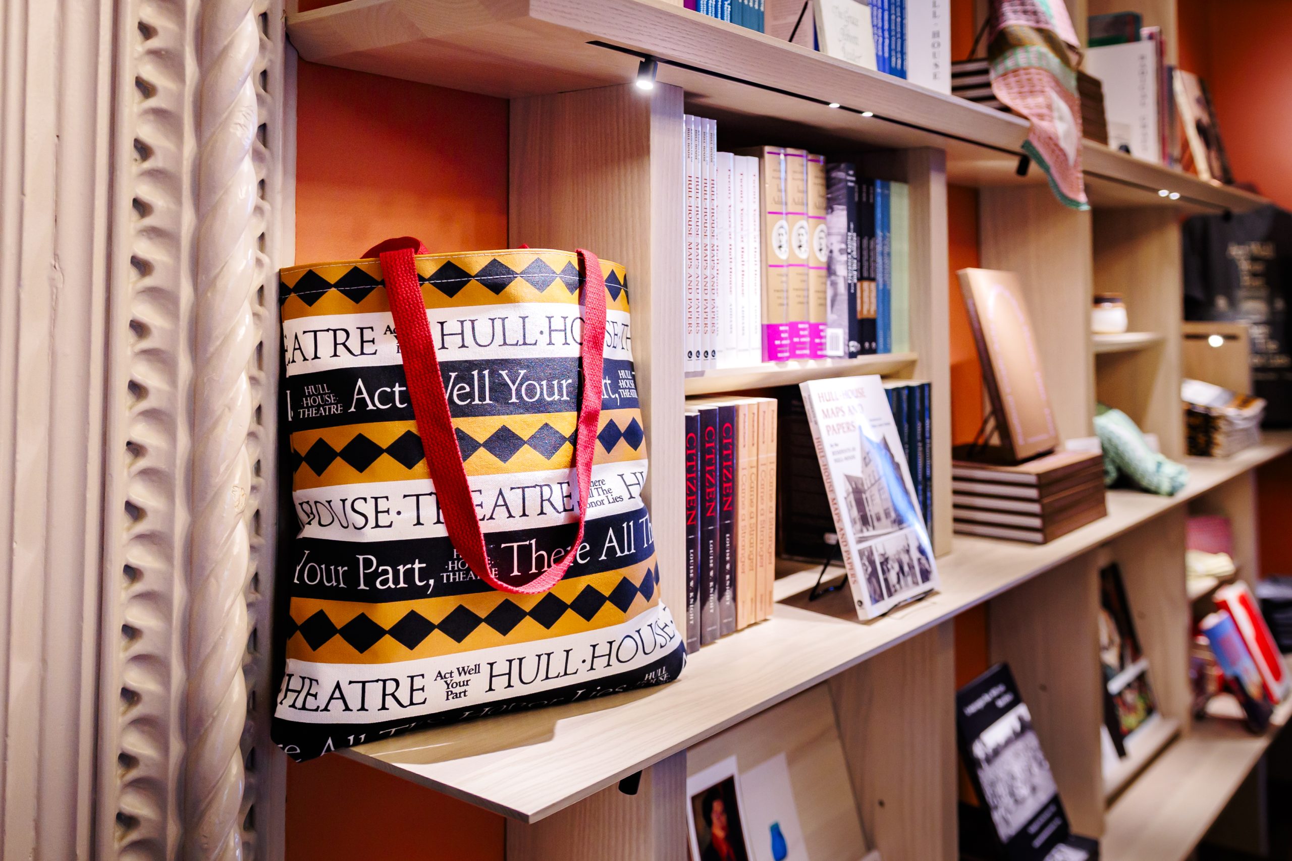A tote bag with a red strap and theater-themed text sits on a wooden bookshelf filled with books and various items in a well-lit store or library.