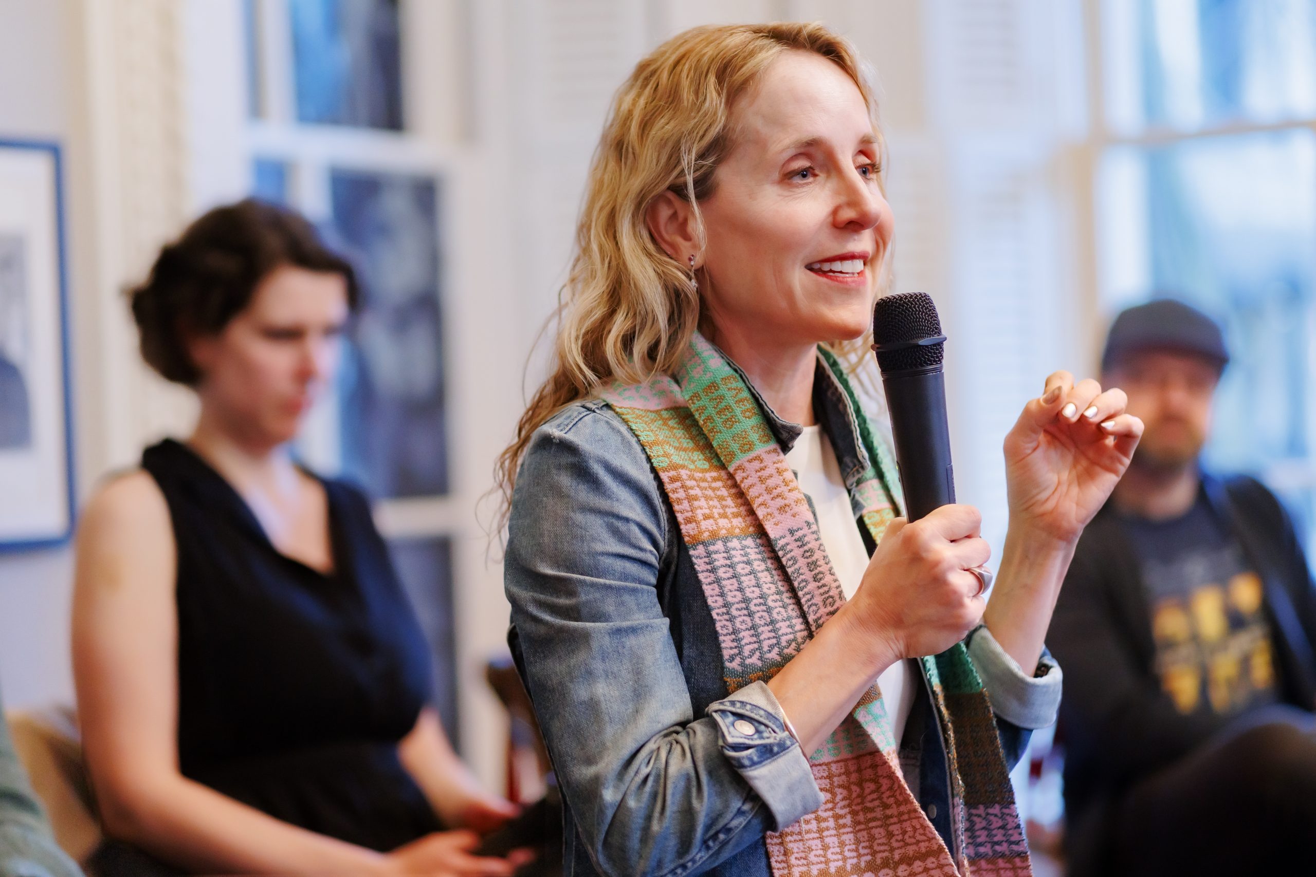 A woman with wavy blonde hair speaks into a microphone, smiling, while wearing a colorful scarf and denim jacket. In the background, two people sit and listen attentively in a bright room.