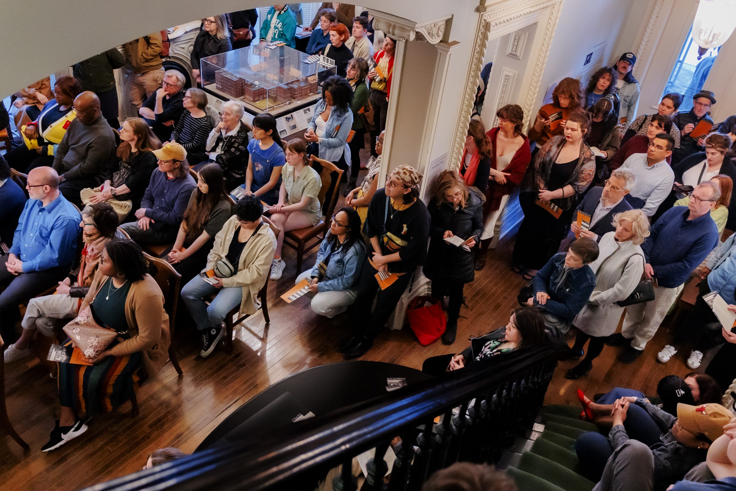 A diverse group of people, some seated and some standing, gather closely together in a well-lit room with wooden floors and ornate walls, viewed from above near a staircase.
