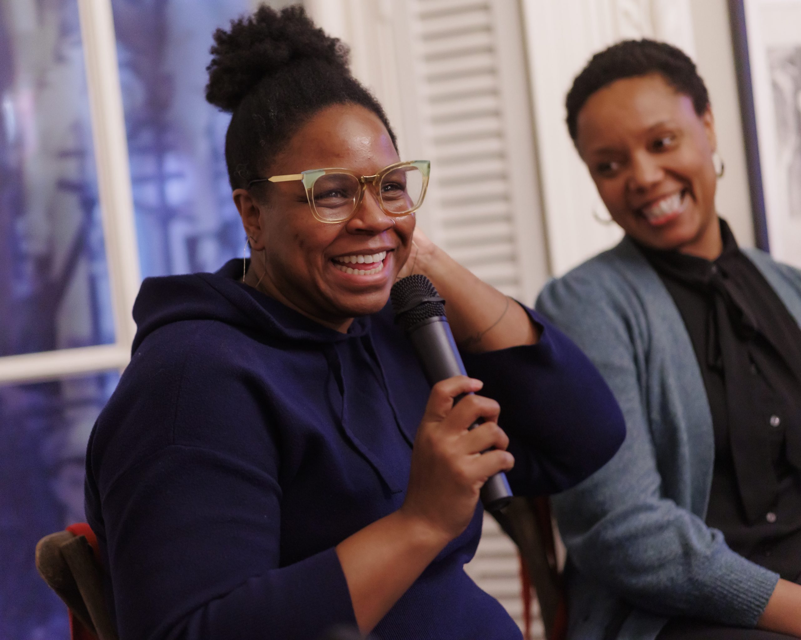 Two women sit next to each other indoors. One woman, smiling and wearing glasses and a navy blue top, holds a microphone while the other woman, also smiling, listens attentively.