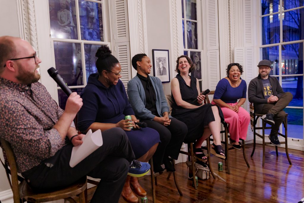 A group of six people sit on stools in a well-lit room with large windows, laughing and talking. One person on the left holds a microphone and paper, leading the panel discussion.