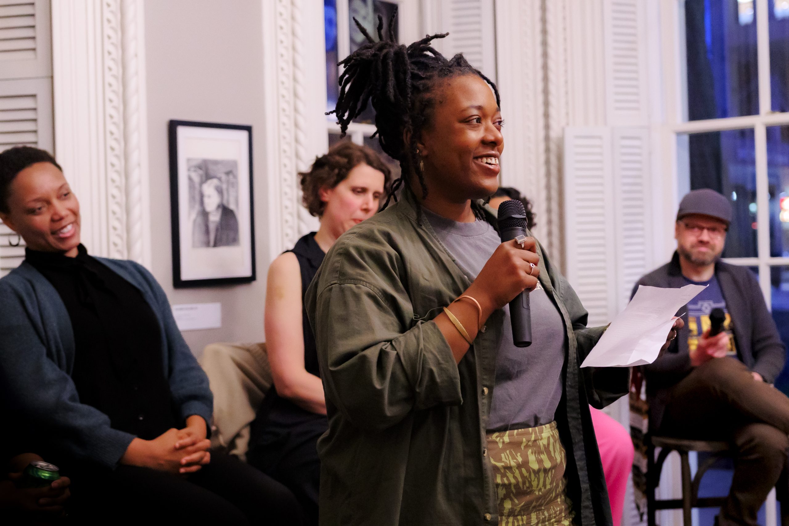 A person smiles while speaking into a microphone and holding a paper, addressing a seated audience in a well-lit room with framed art and large windows. Four people are seated and listening attentively.