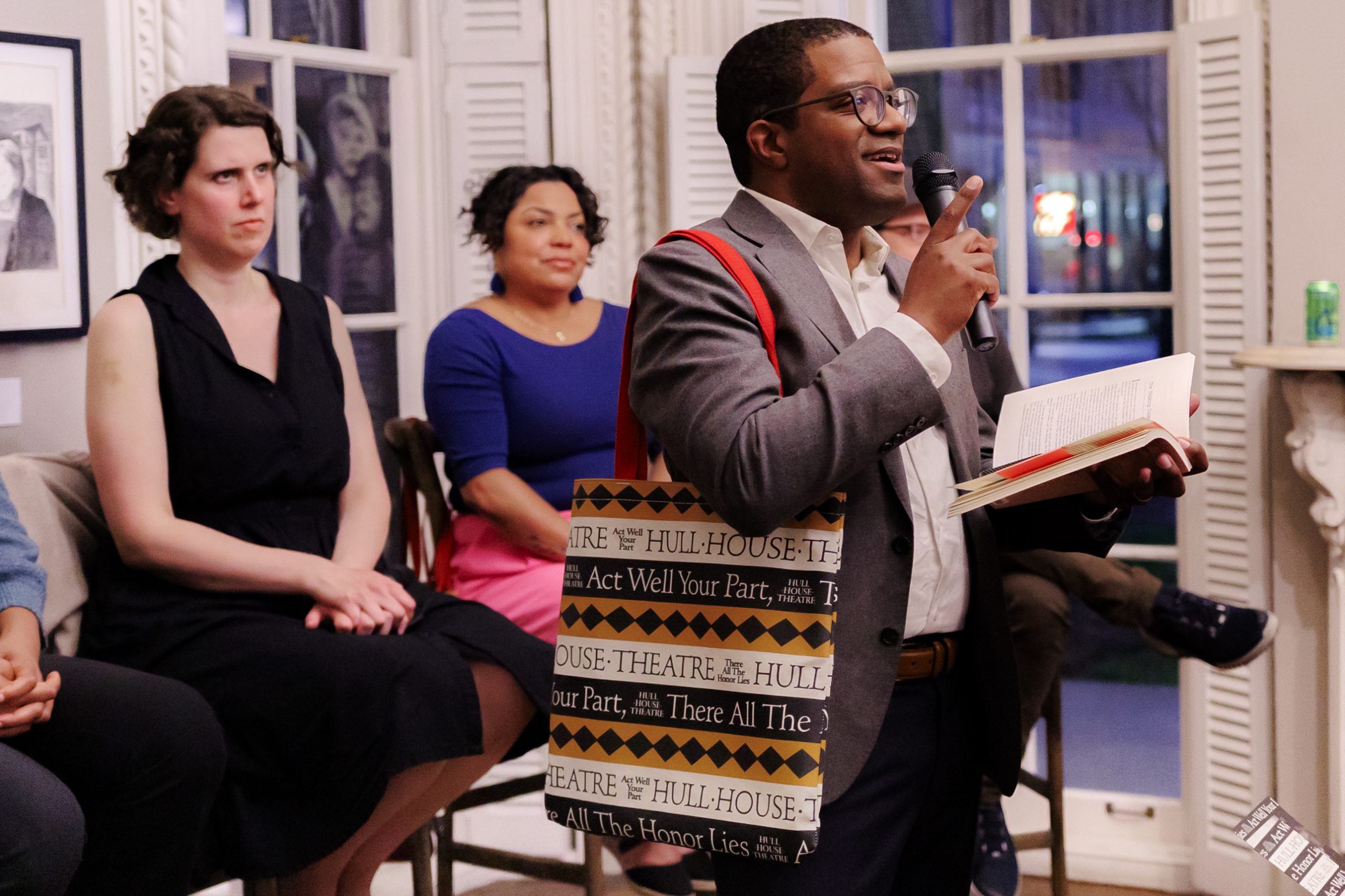A man holding a microphone and an open book speaks to an audience. He wears glasses, a gray blazer, and a patterned tote bag. Four people are seated behind him, listening attentively in a room with white walls and open shutters.