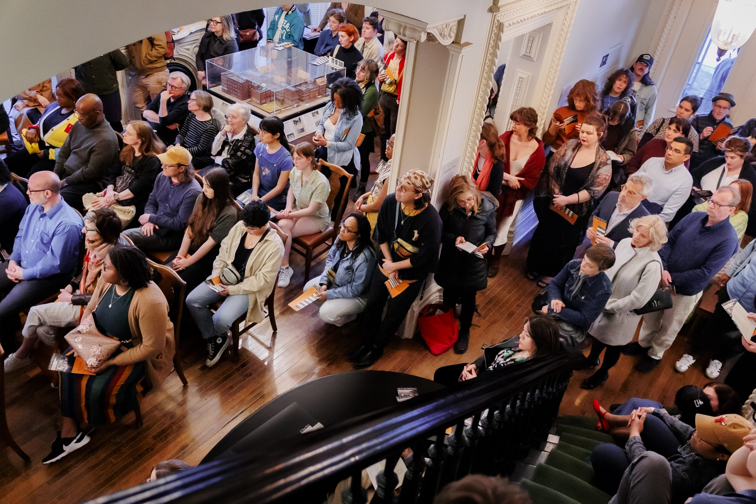 A large, diverse group of people, seated and standing, gather indoors for an event. The view is from above near a staircase. Attendees appear attentive, holding programs or booklets in a well-lit, ornate room.
