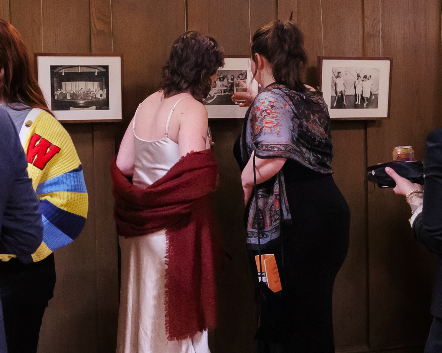 Two women stand closely together, facing framed black-and-white photos on a wooden wall. One wears a white dress and red shawl, the other a black outfit with a patterned shawl. Other people are partially visible nearby.