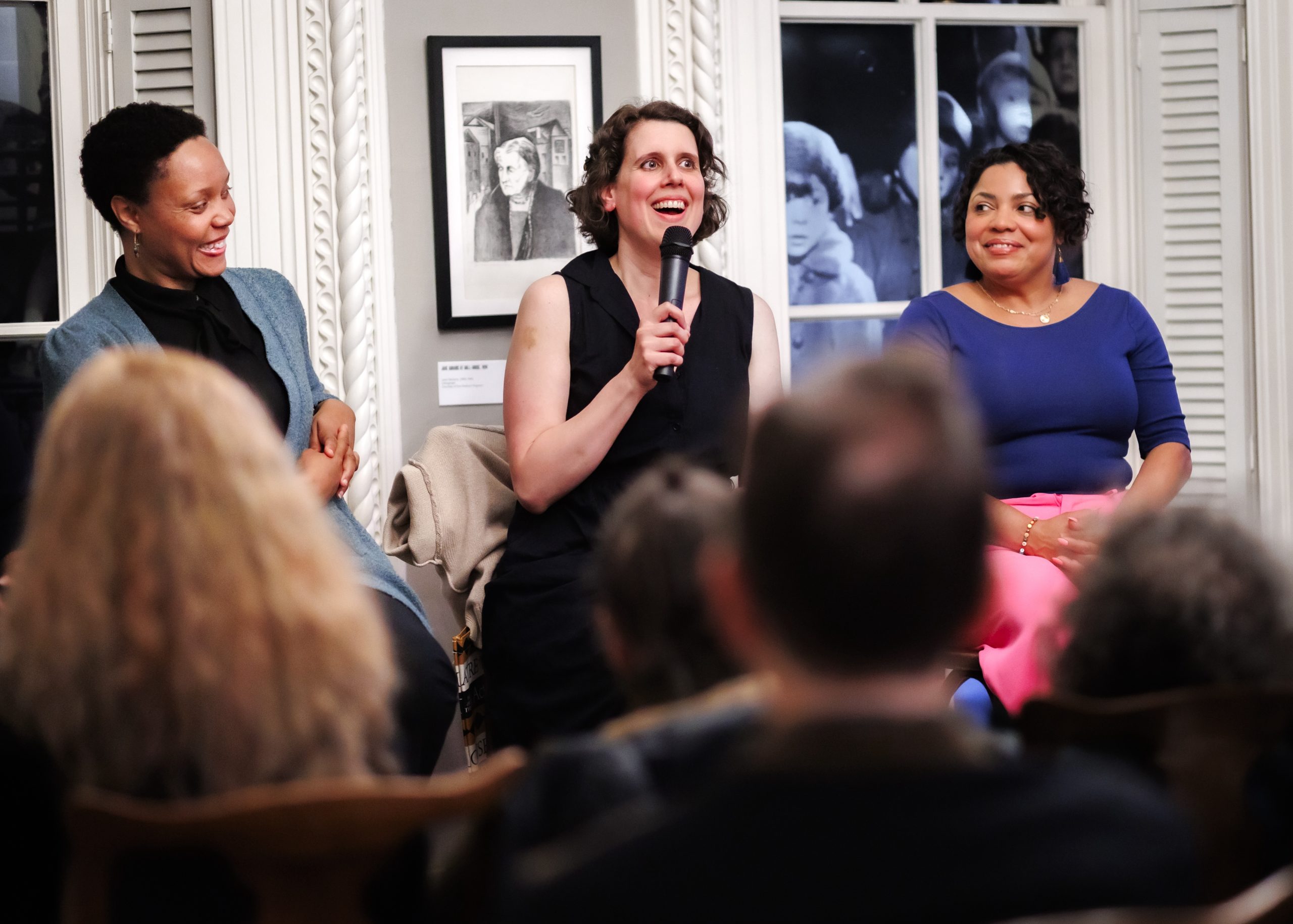 Three women sit in front of an audience, engaged in a panel discussion. The woman in the center is smiling and speaking into a microphone, while the other two women sit on either side, listening and smiling.