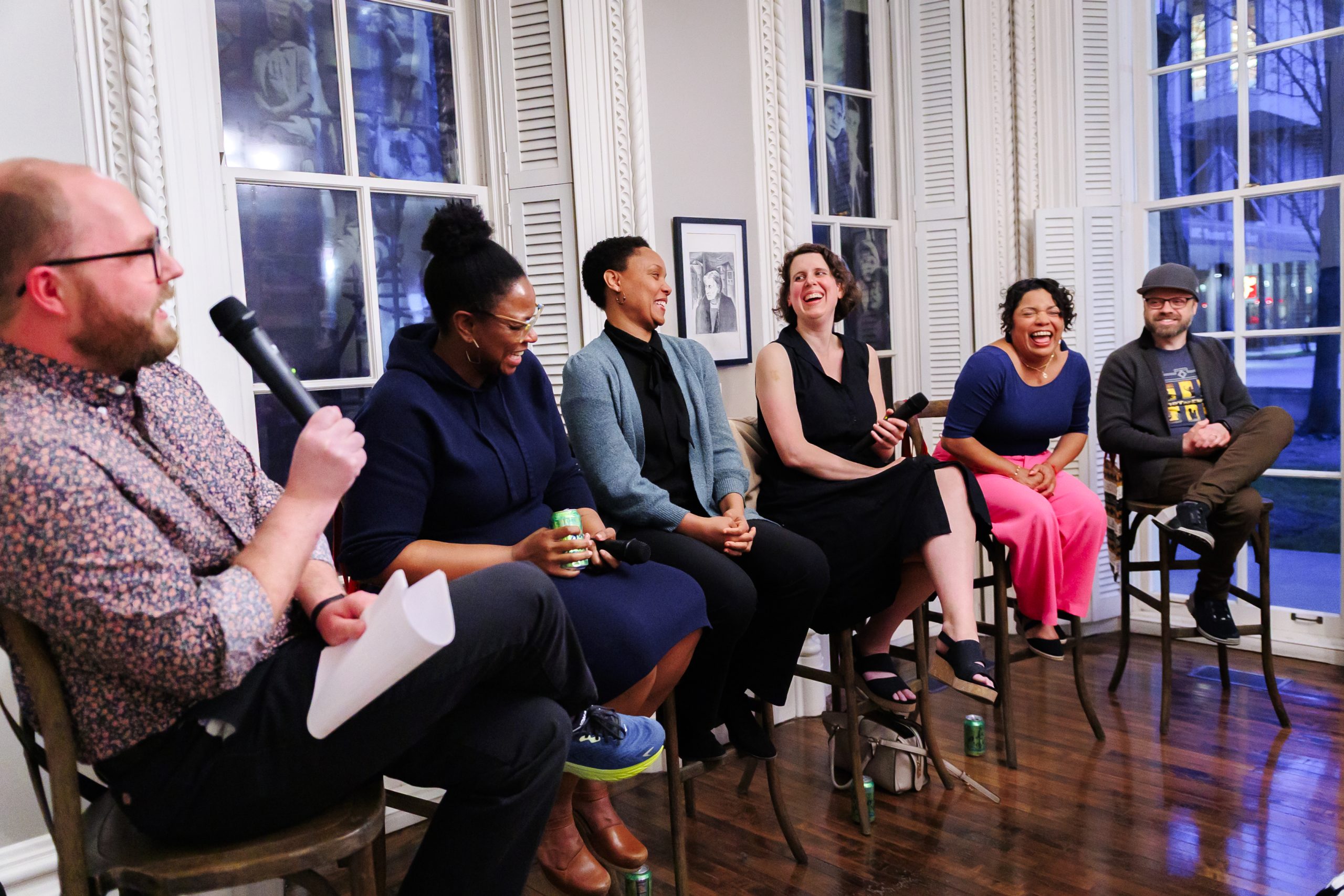 Six people sit on stools in front of large windows, laughing and talking. One person on the left holds a microphone and papers, leading the discussion. The atmosphere is relaxed and engaging.
