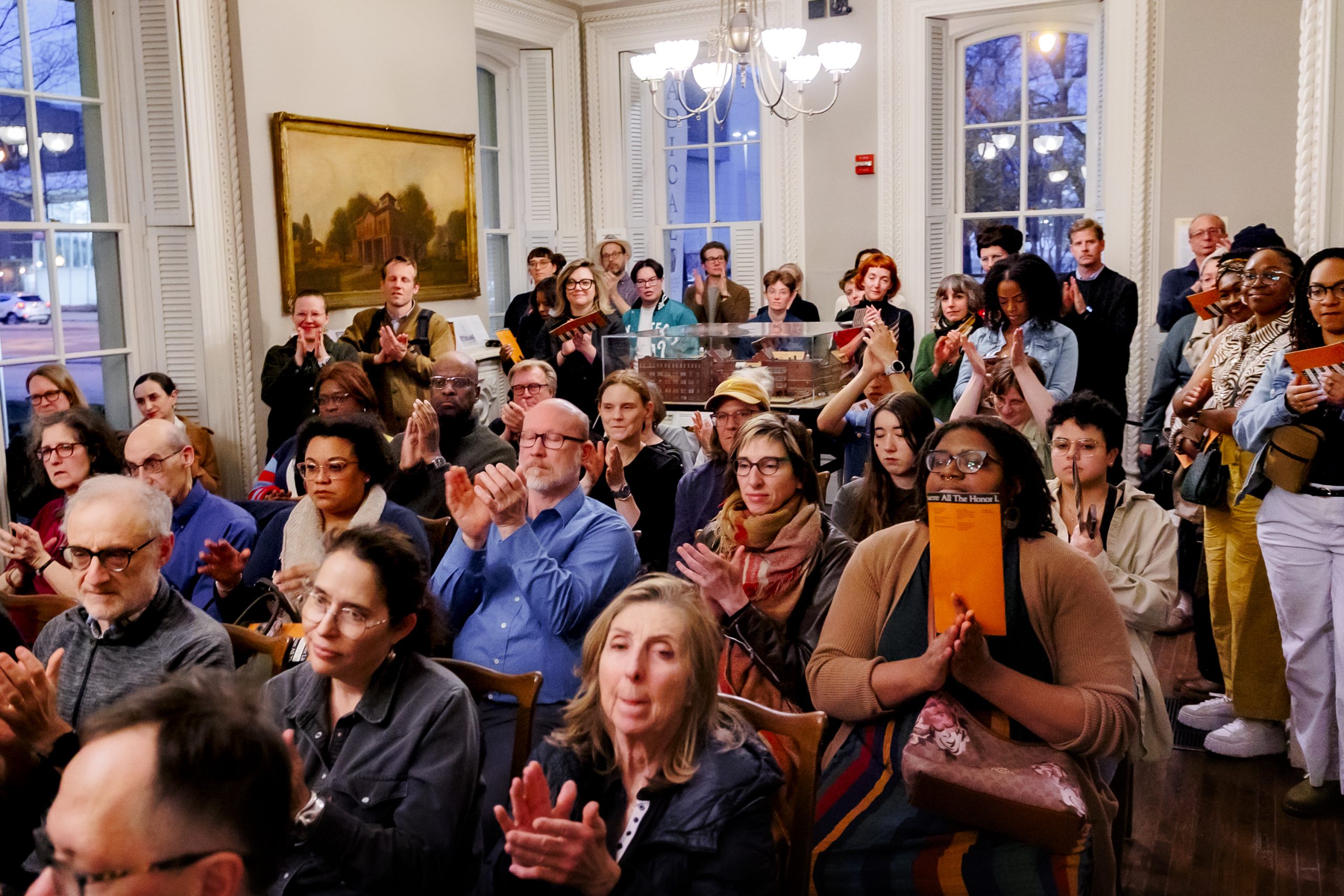 A diverse group of people sit and stand in a well-lit room, attentively watching an event and applauding. The room has large windows, white walls, and a painting on the wall.
