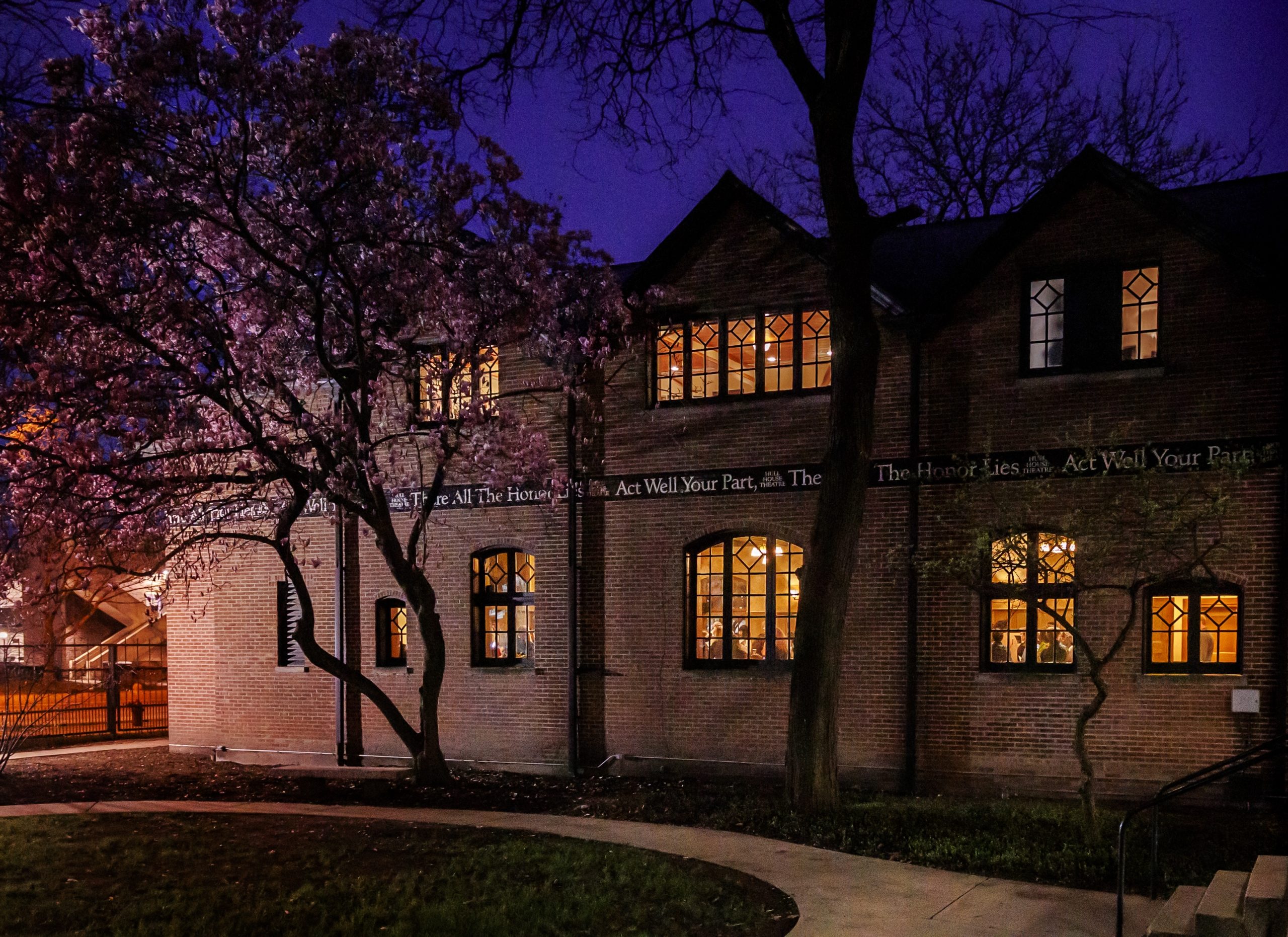 A brick building glows warmly with light from its windows at dusk. Flowering trees with pink blossoms stand in front, and a path curves through a grassy area. The sky is a deep blue, creating a peaceful evening scene.