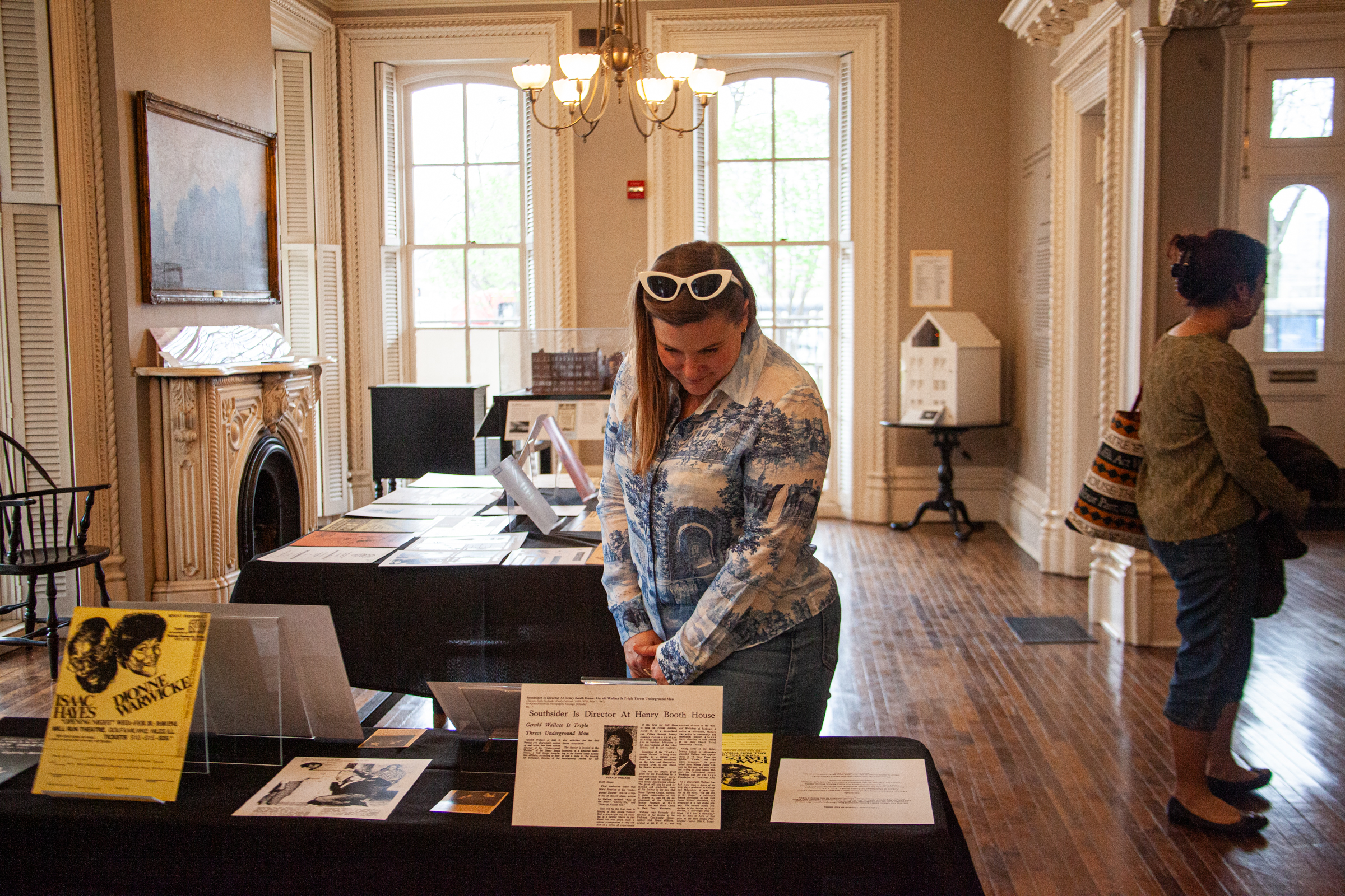 A woman in a blue and white shirt reads a display on a table in a bright, elegant room with large windows and wood floors. Another person stands nearby, looking at exhibits arranged throughout the space.