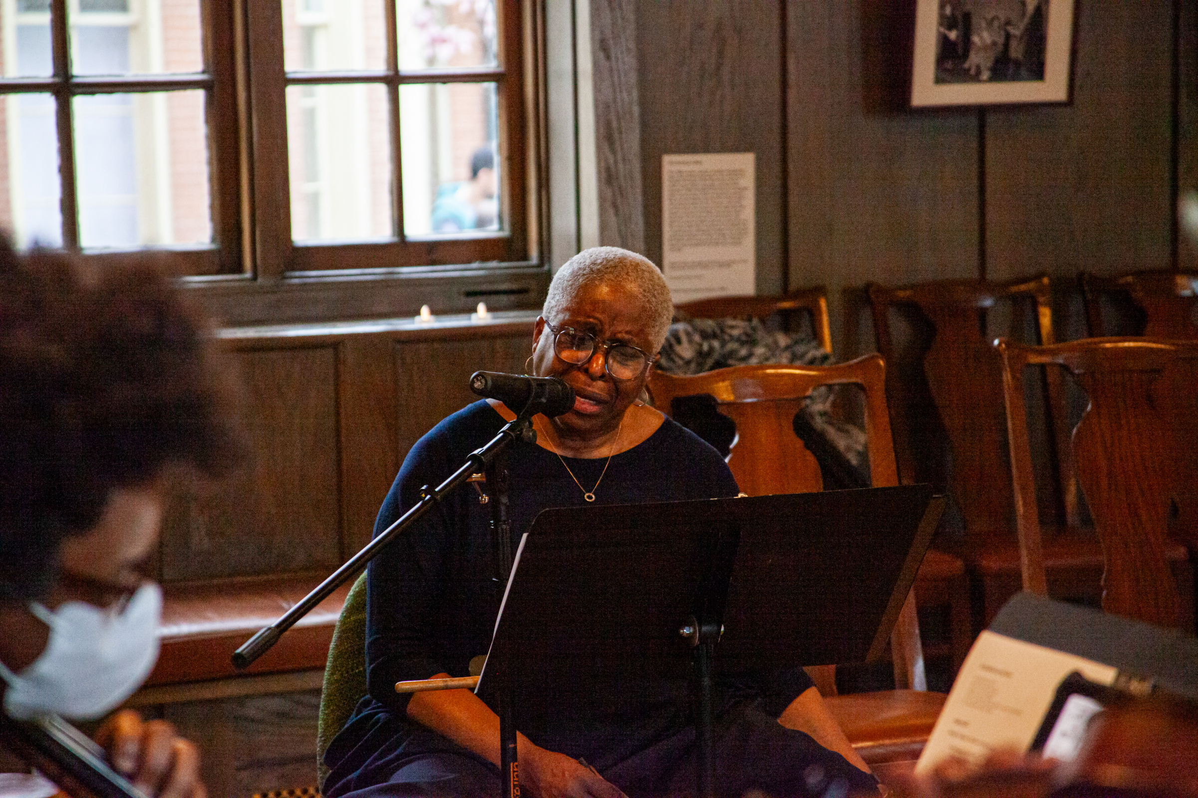 An older woman with short gray hair and glasses sits at a music stand, speaking or singing into a microphone in a wood-paneled room. Other musicians and empty chairs are visible in the background.