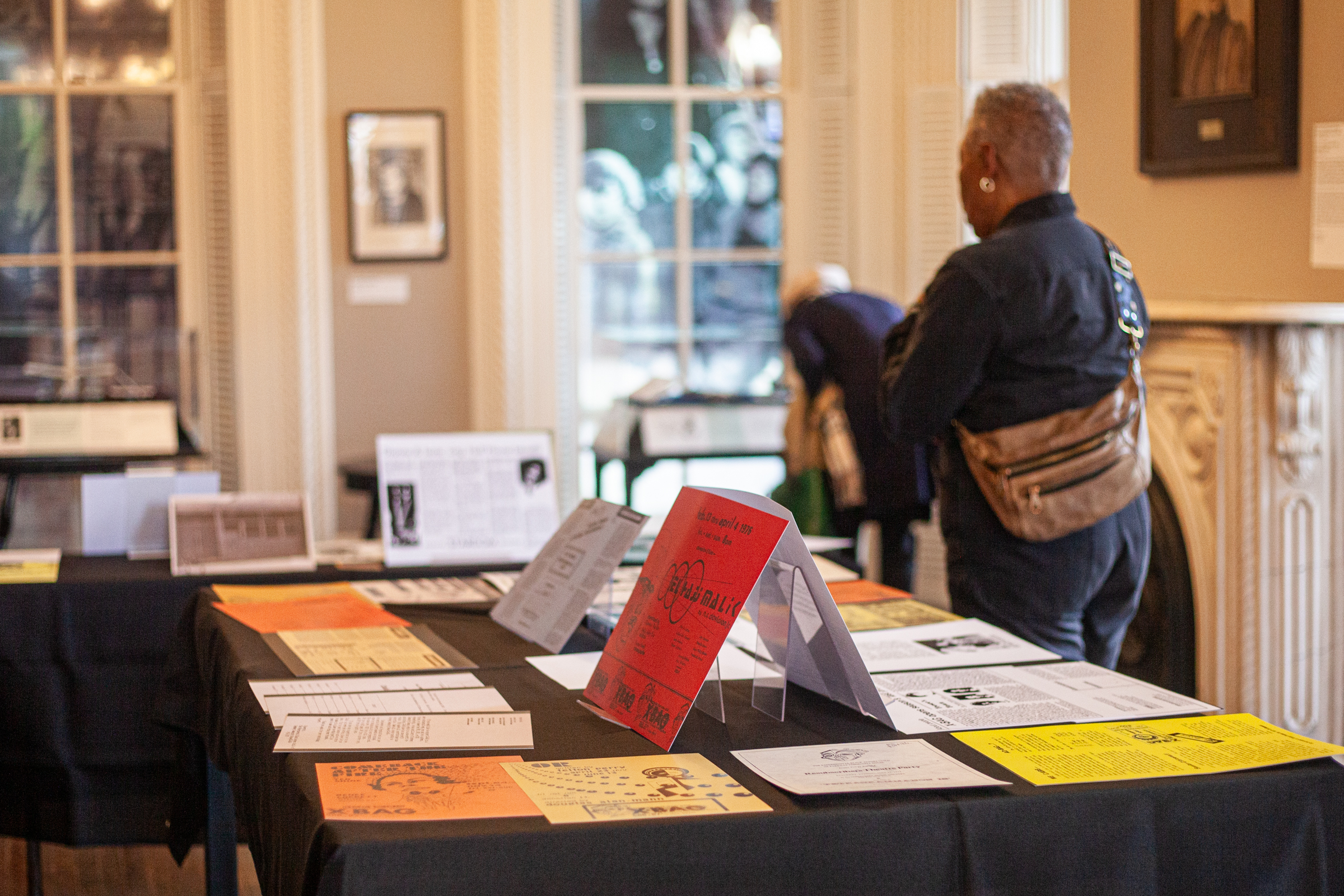 Two people view an exhibit featuring colorful papers, pamphlets, and booklets displayed on tables in a well-lit room with large windows and framed artwork on the walls.