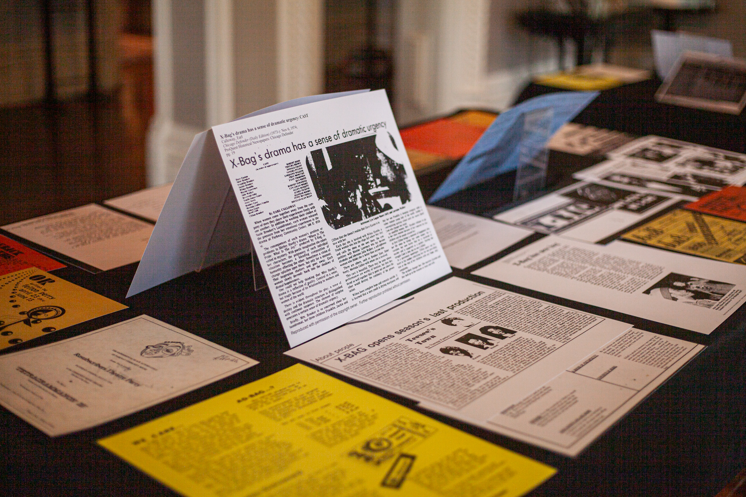 A display table with various printed documents, articles, and pamphlets arranged neatly, including a prominent black-and-white newspaper clipping placed upright among colorful papers.