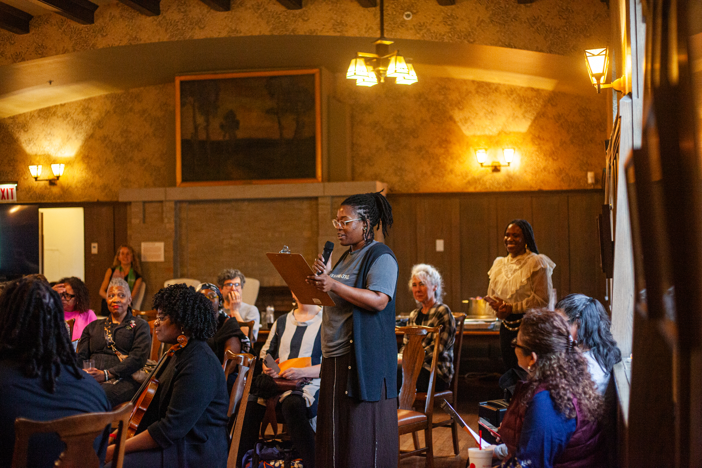 A woman stands and speaks into a microphone while holding a clipboard in a warmly lit room filled with seated people, some of whom are smiling and listening attentively.