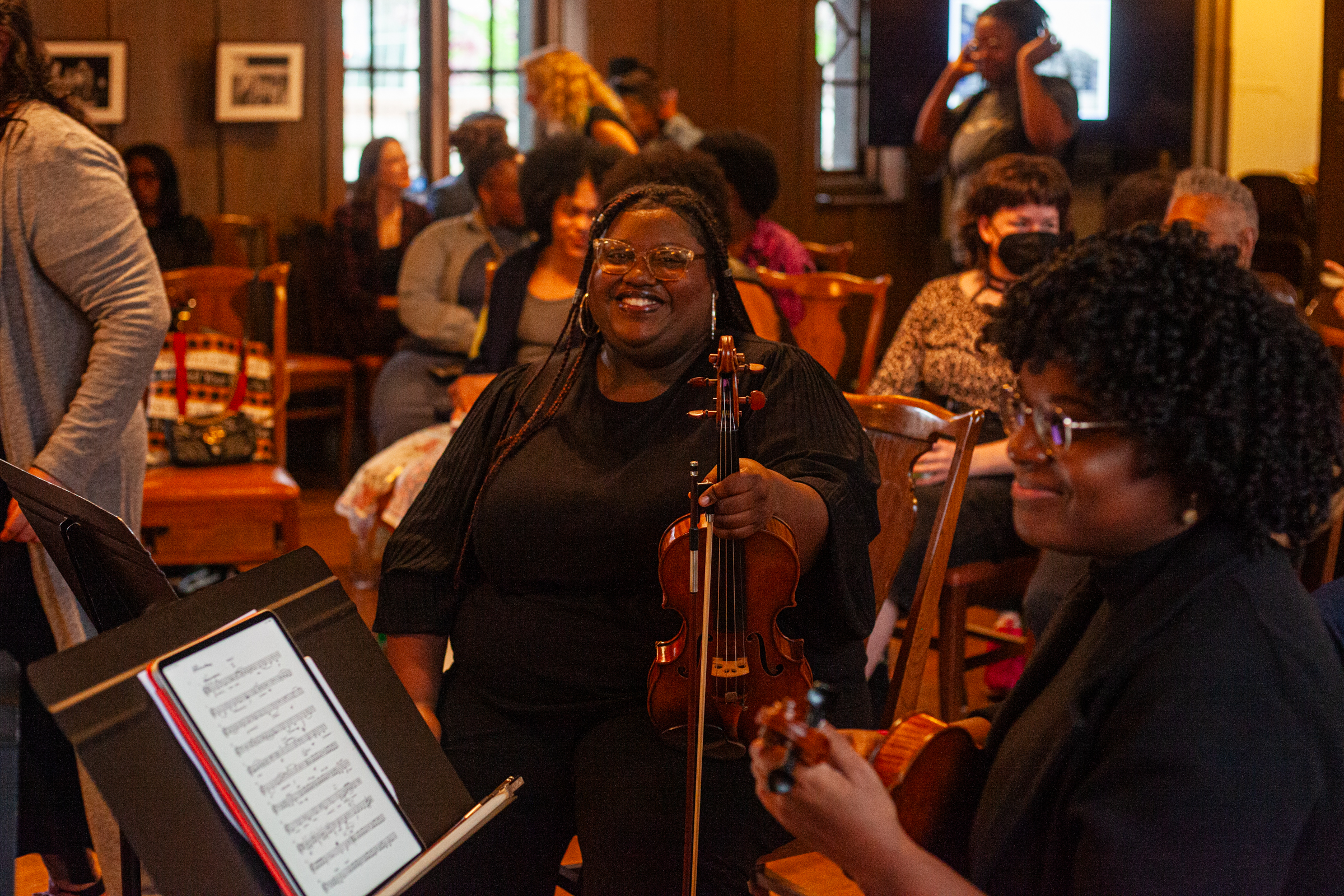 A woman holding a violin smiles while seated among other musicians and attendees in a warmly lit room. Sheet music is visible in the foreground, and people are engaged and interacting in the background.