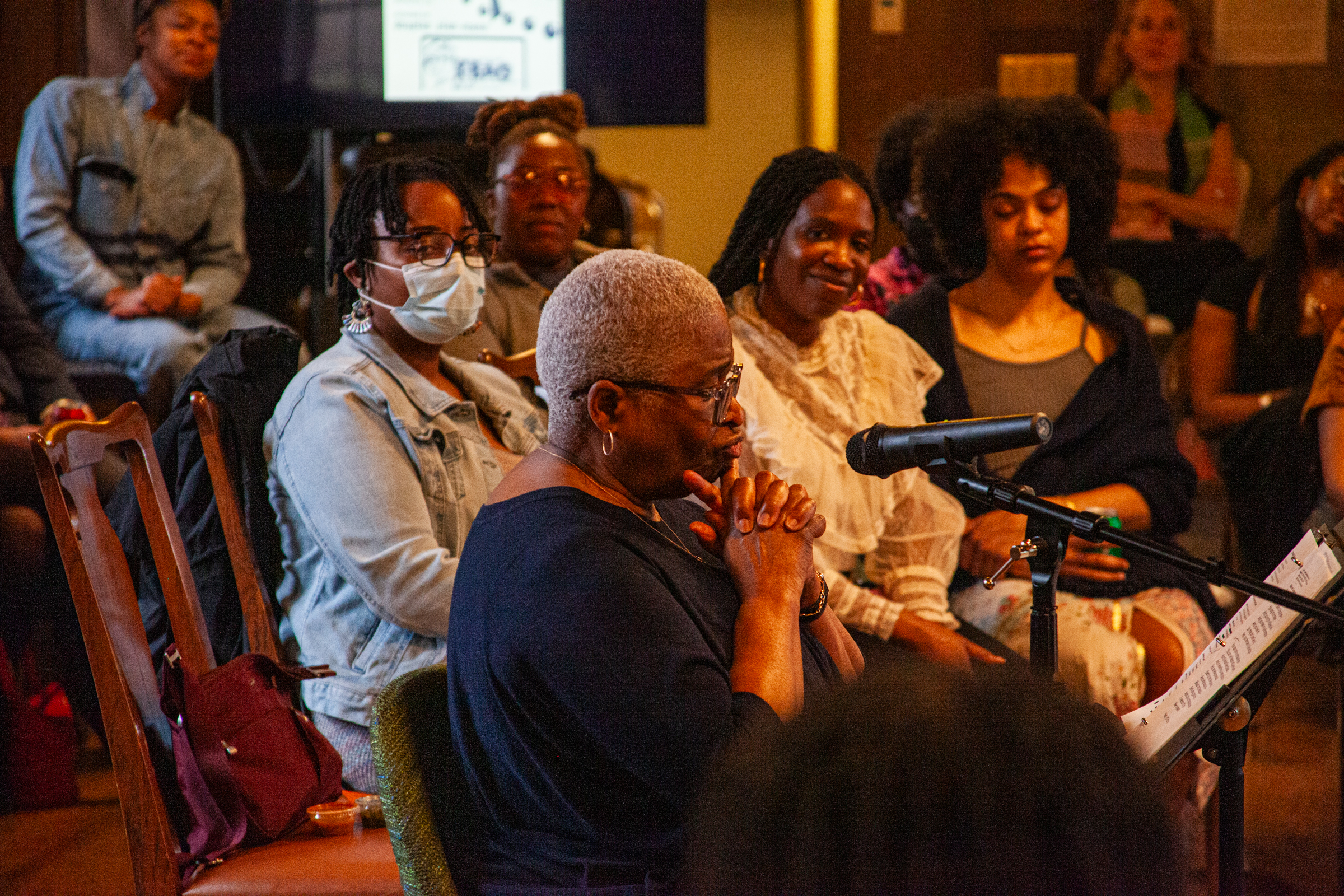 A woman with short gray hair speaks into a microphone, reading from papers, while a diverse audience of women listens attentively in a warmly lit room.