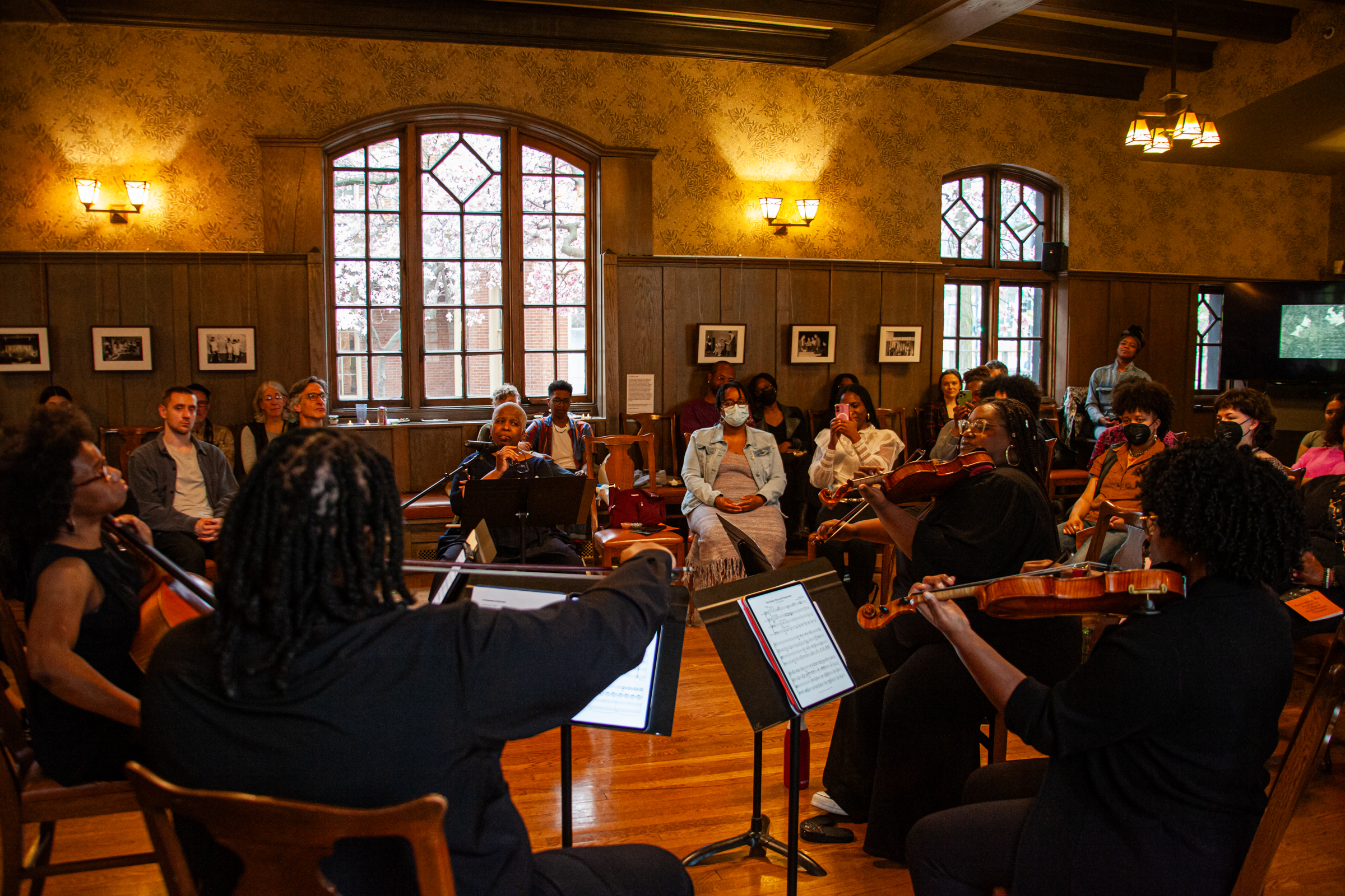 A string quartet performs for an audience seated in a warmly lit, wood-paneled room with large windows, framed photos on the walls, and a polished wooden floor. Some audience members wear masks.