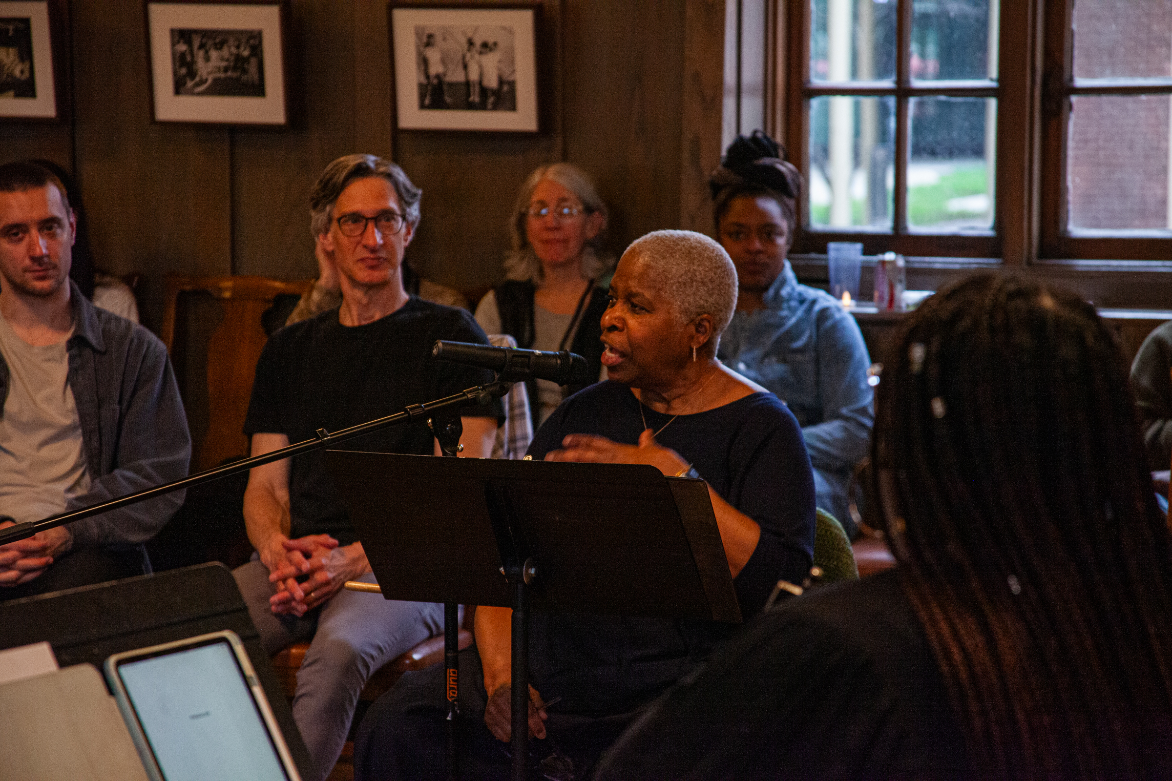 An older woman speaks into a microphone while seated indoors, surrounded by an attentive audience. Framed photos hang on the wood-paneled wall behind them, and daylight streams through large windows.