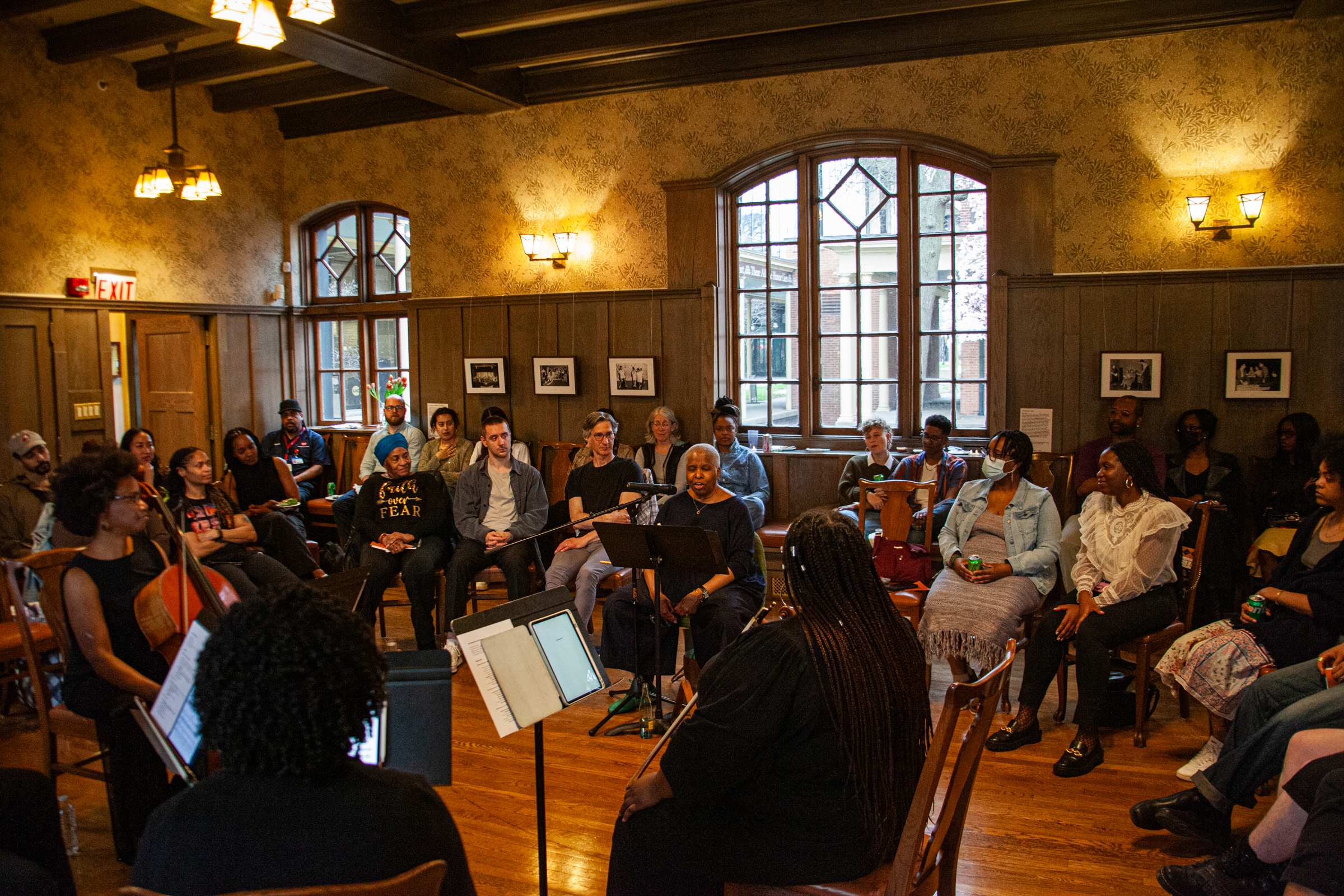 A diverse group of people sits in a warmly lit room, arranged in a circle, listening attentively to speakers and musicians at the center. Framed photos decorate the wood-paneled walls, and natural light streams through large windows.