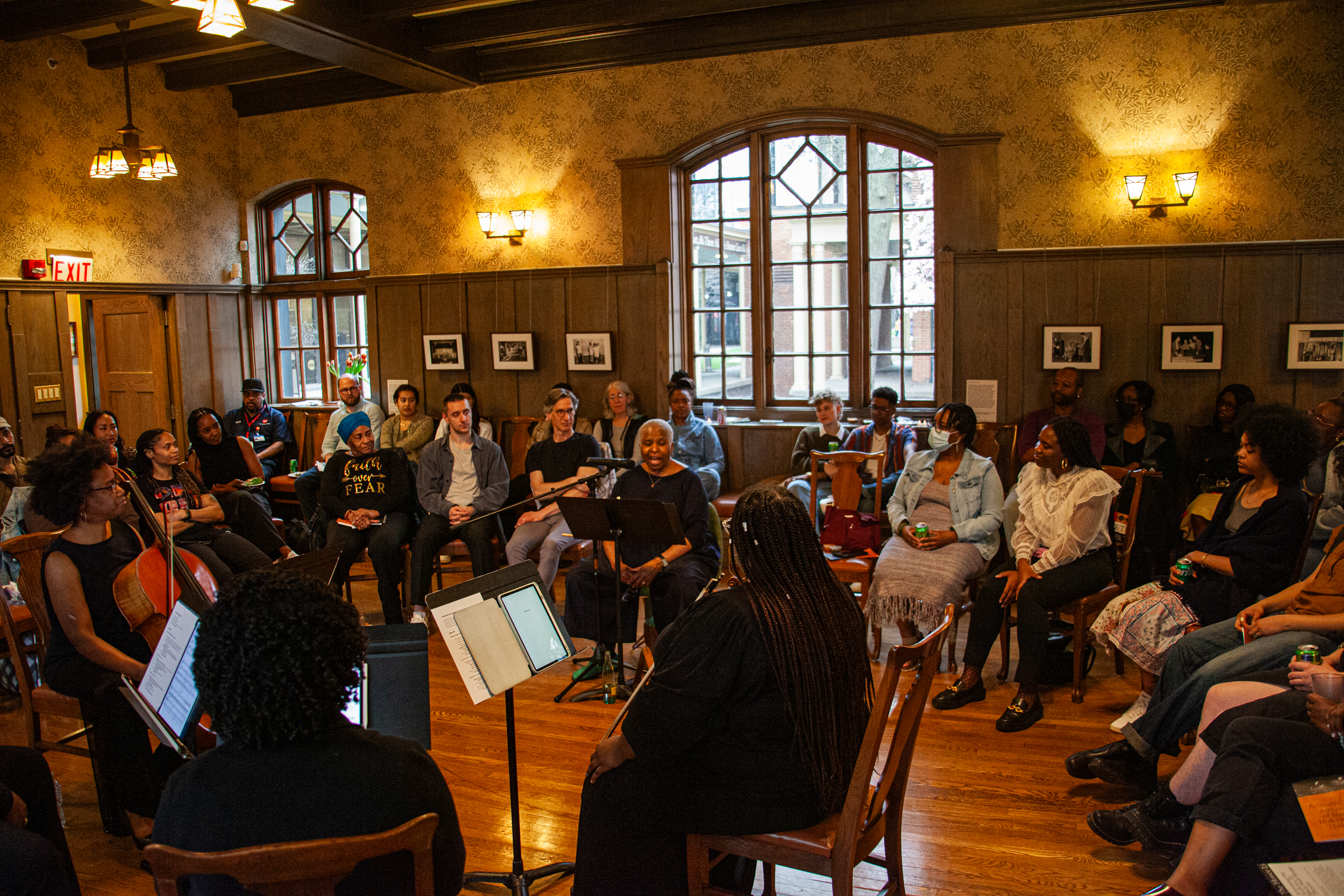 A diverse group of people sit in a circle in a warmly lit room with wooden floors and walls, listening to musicians performing with string instruments in the center. Framed photos hang on the walls behind them.