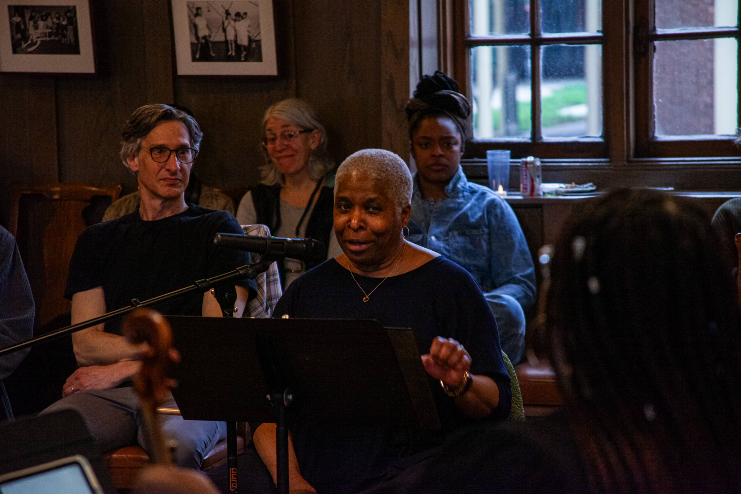 A woman speaks into a microphone while seated at a music stand, surrounded by several people in a warmly lit room with wooden walls and large windows in the background.