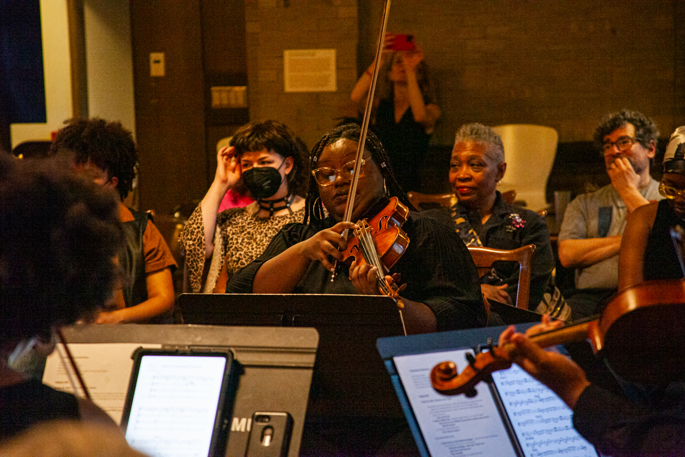 A violinist plays in an orchestra, focused on her music, while audience members watch attentively; some are smiling, others wear masks, and sheet music is visible in the foreground.