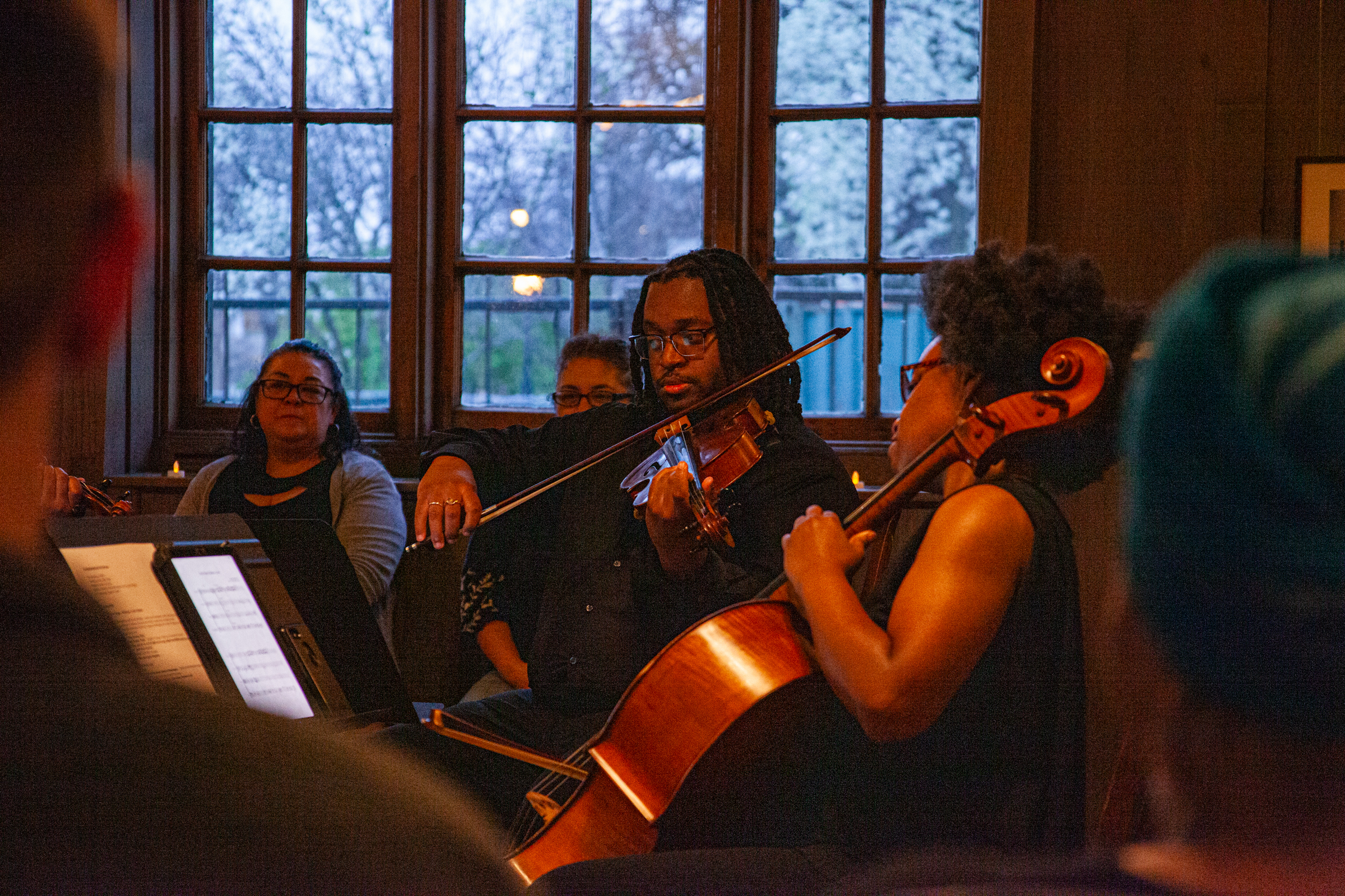 Four musicians perform indoors near large windows; one plays a violin, another a cello, while two others watch. Warm lighting and evening scenery outside create an intimate atmosphere.