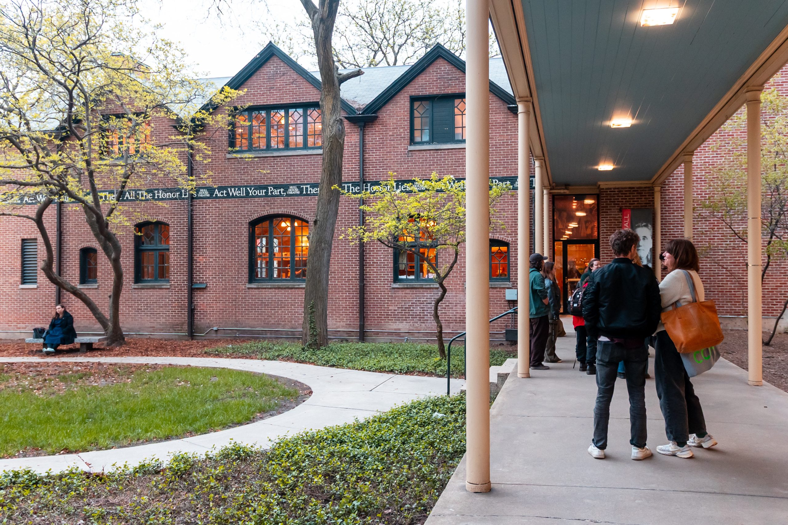 People stand and chat under a covered walkway outside a brick building with large windows, surrounded by trees and a grass courtyard. Some people are near the entrance, and others sit on the low wall in the courtyard.