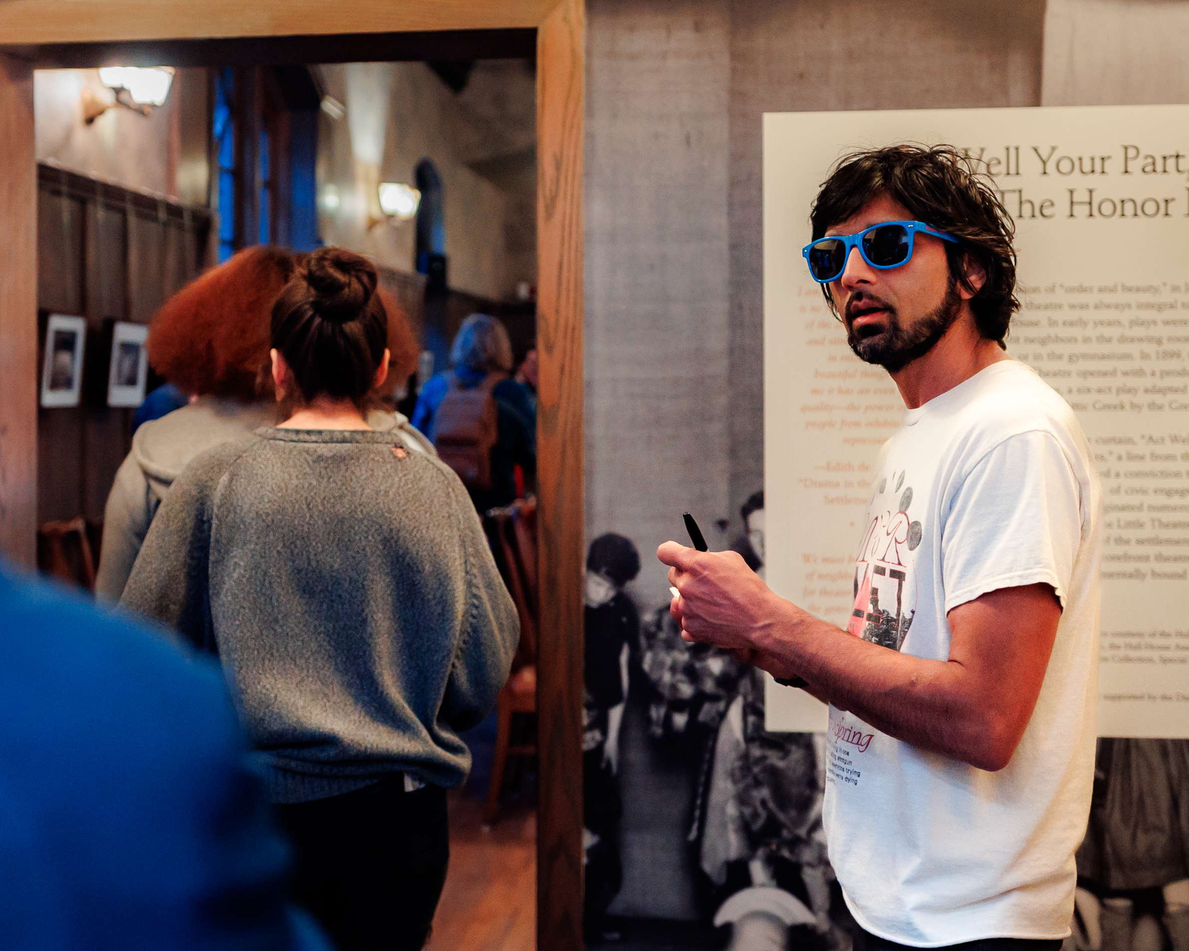 A person with dark hair, a beard, and blue sunglasses holds a pen and paper in an indoor space with framed photos on the walls, while several people stand in the background facing away.