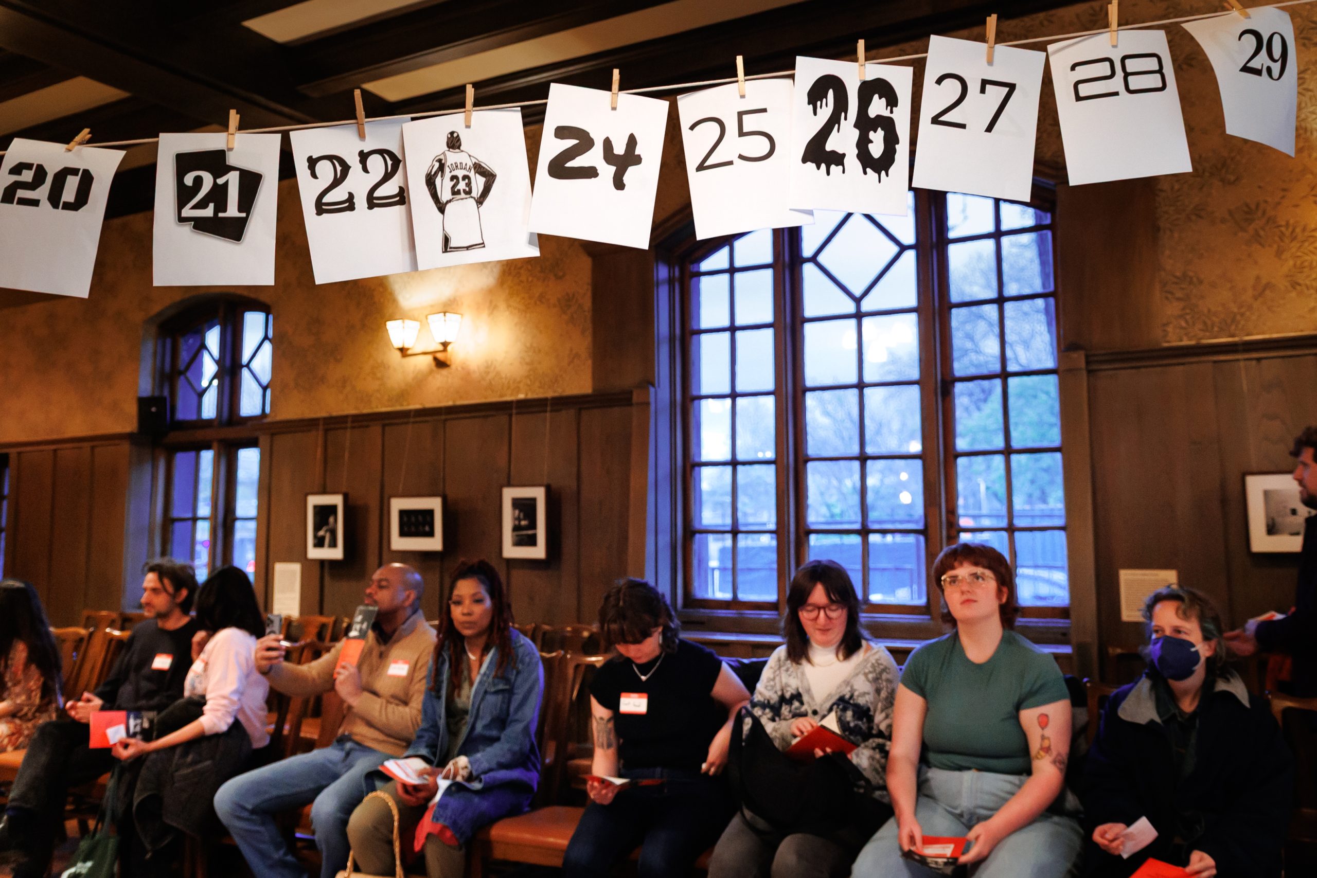 A group of people sit in a wood-paneled room under hanging paper signs displaying jersey numbers 20 to 29, each with a different design. Large windows and framed photos are visible in the background.