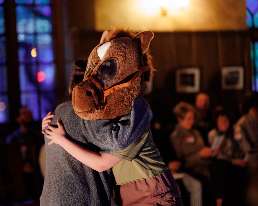 Two people hug indoors; one wears a large, cartoonish horse head mask. Several people sit in the background, watching the scene in a warmly lit room with wooden paneled walls and framed photos.
