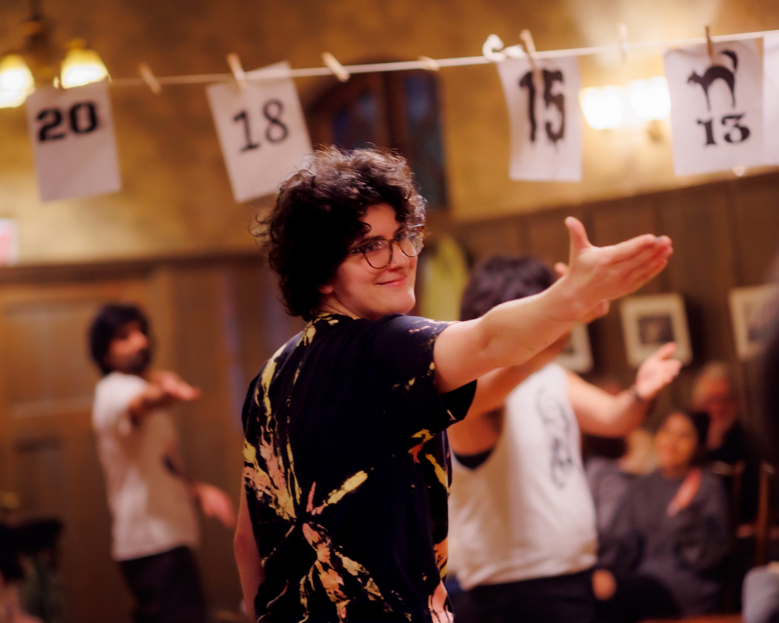 A person with curly hair and glasses extends their arm forward, smiling. Other people are in the background, and numbered signs (20, 18, 15) hang on a line above them in a warmly lit indoor space.