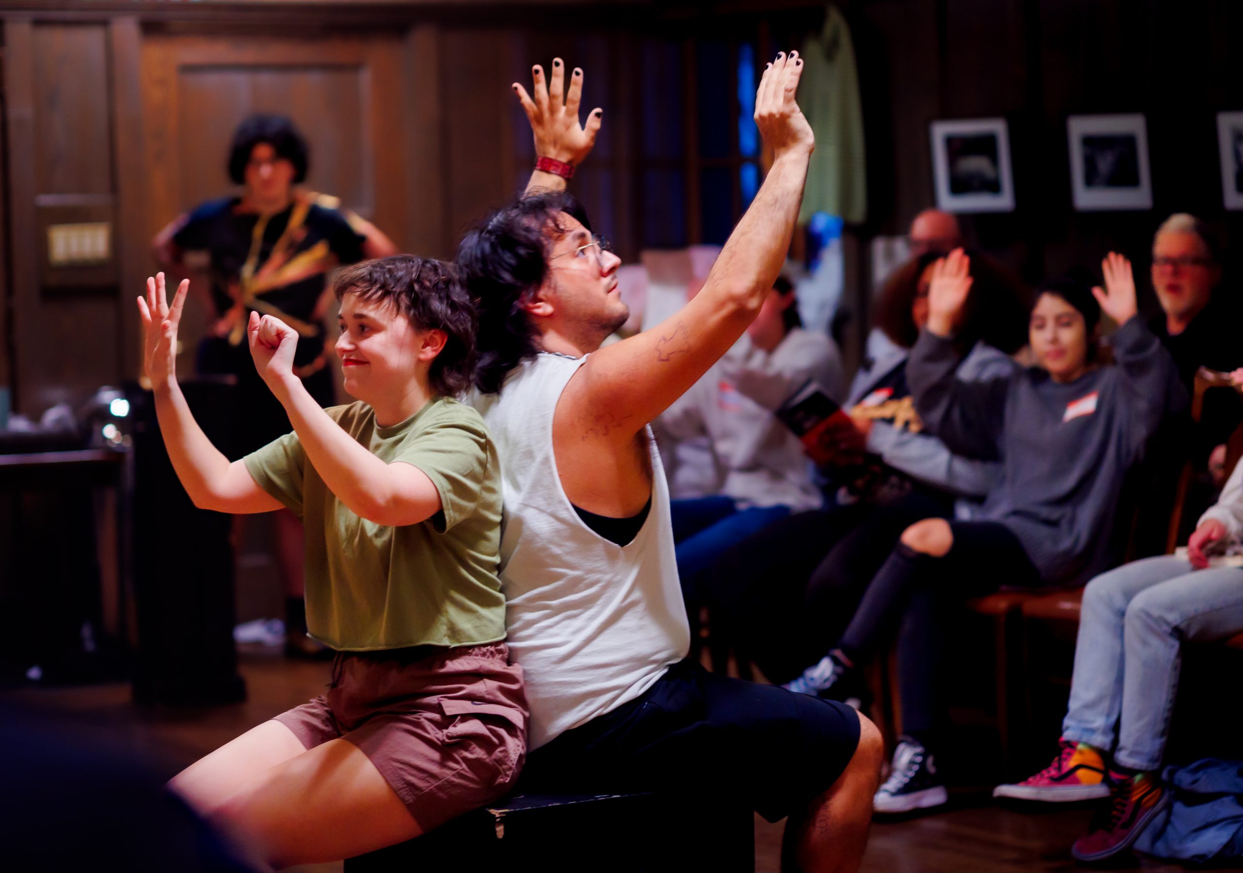 Two people sit back to back on a bench, raising their arms with expressive hands, while others in the background also raise their hands, suggesting a performance or interactive workshop in a room with wooden walls.