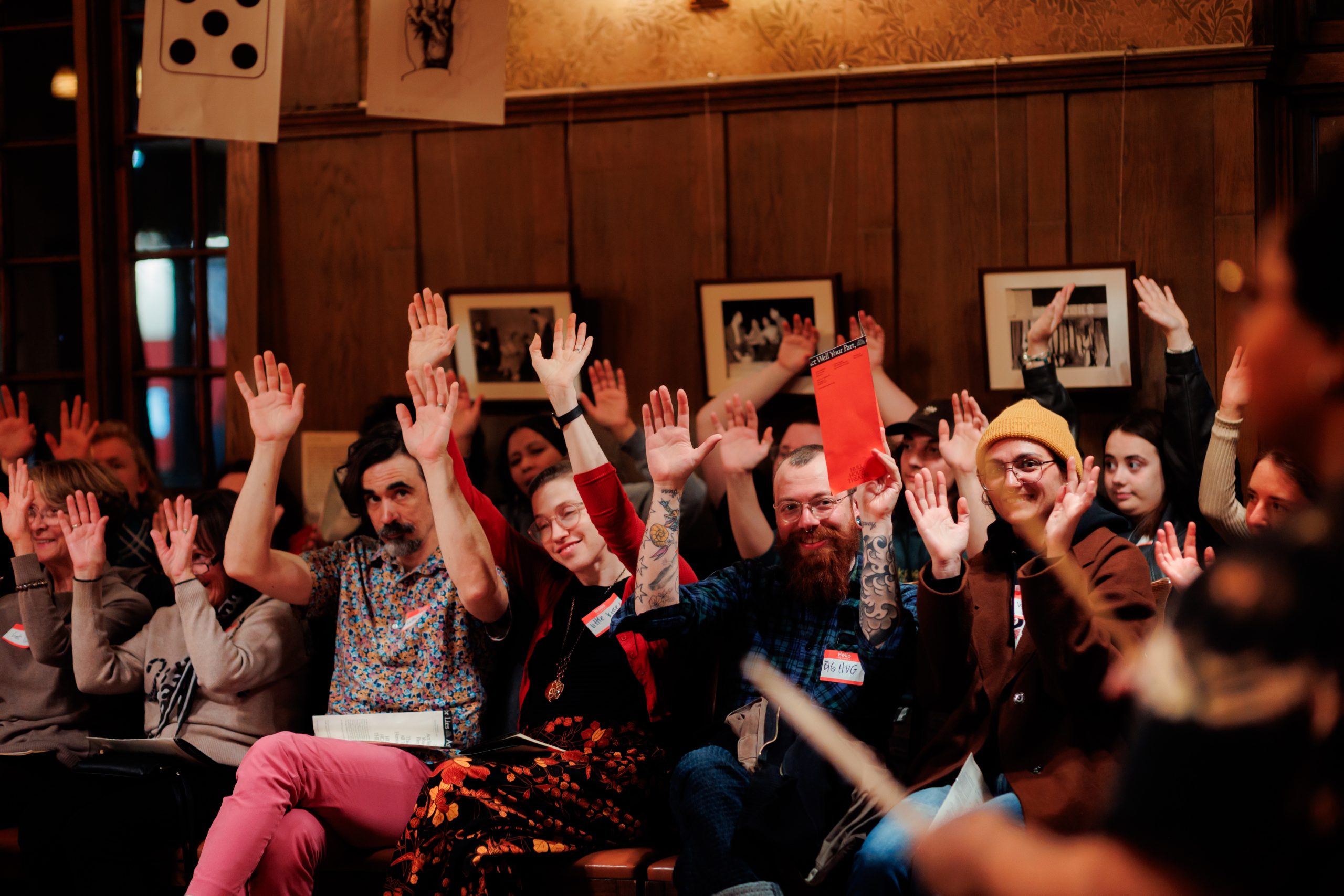 A group of people sitting indoors, raising their hands enthusiastically, as if participating in a discussion or vote. The room has wooden walls and framed pictures, creating a warm, inviting atmosphere.