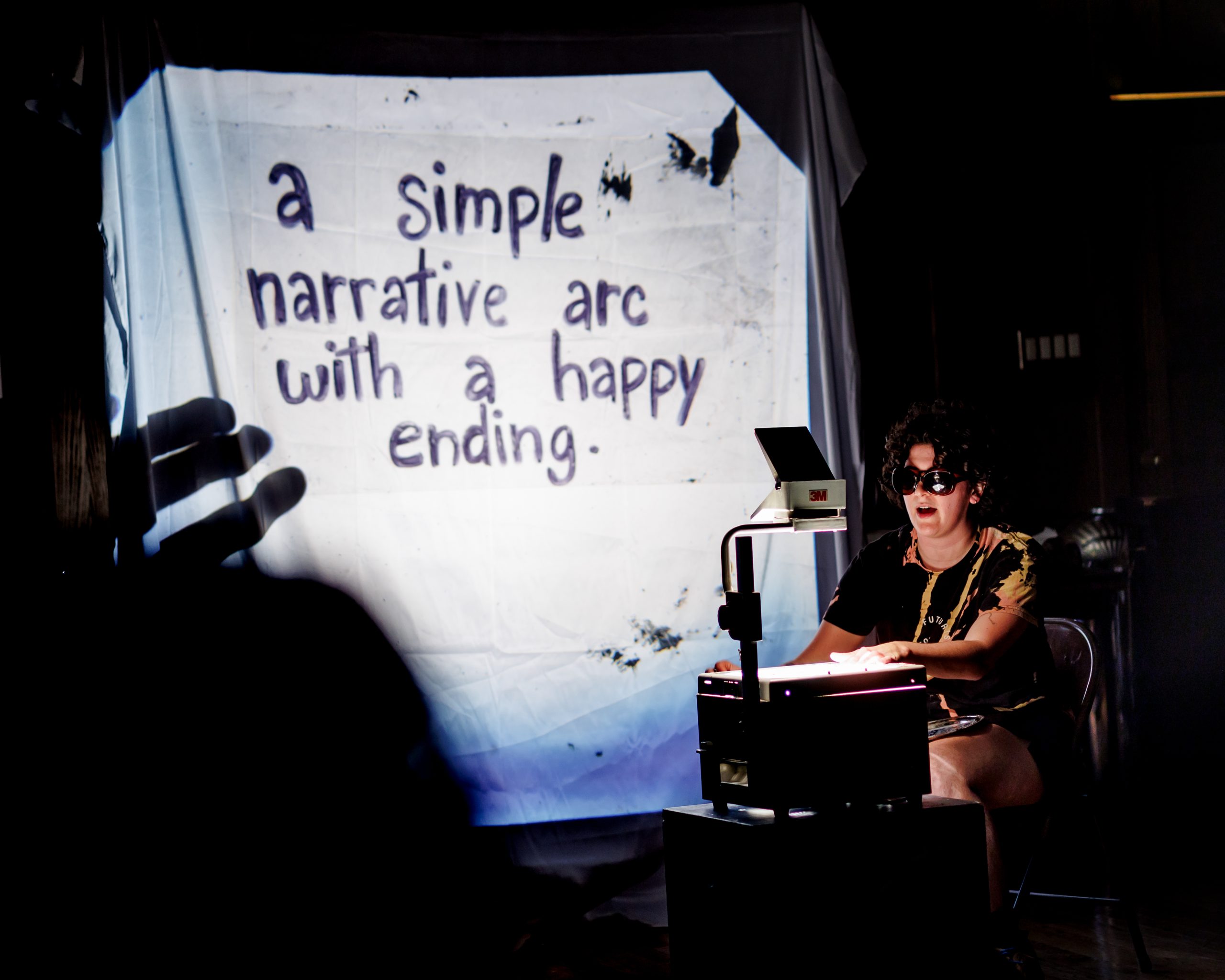 A person in sunglasses sits at an overhead projector, projecting the words a simple narrative arc with a happy ending onto a sheet, with a shadowy hand visible on the projection.