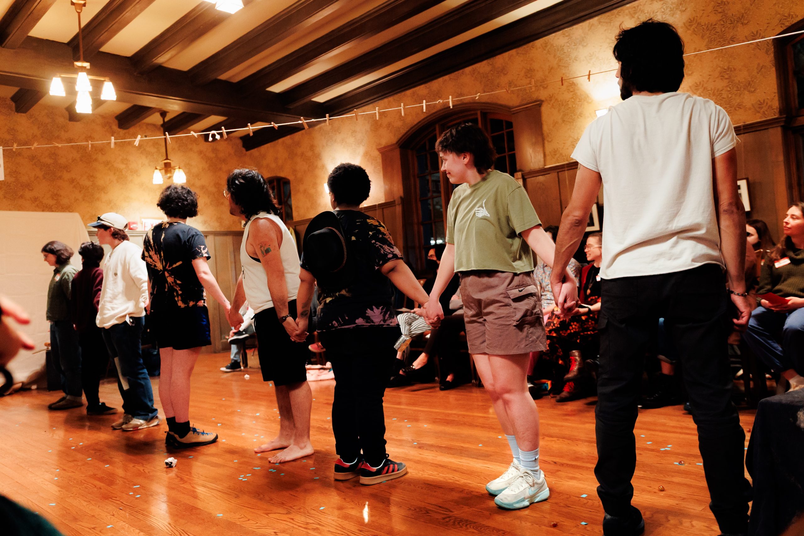 A group of people stand in a line holding hands in a warmly lit room with wooden floors, while others sit and watch from chairs along the wall. The atmosphere appears casual and communal.