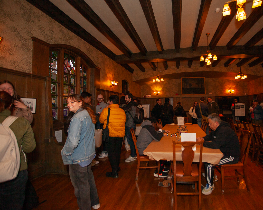A group of people gathers in a warmly lit, wood-paneled room with beamed ceilings. Some are seated at a table, others stand and chat. A woman in a denim jacket smiles at the camera. Stained glass windows are visible.