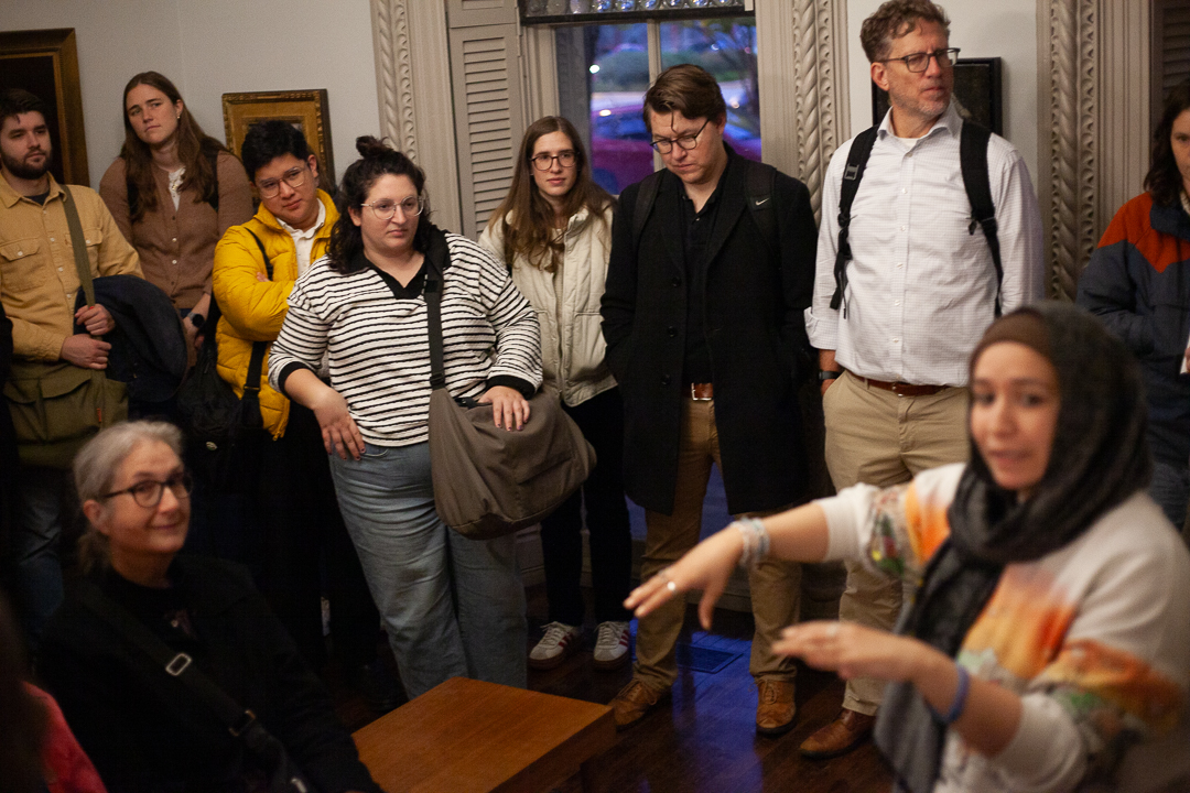 A group of people stand closely together indoors, attentively listening to a woman in the foreground who is gesturing with her hands. The atmosphere appears focused and engaged.