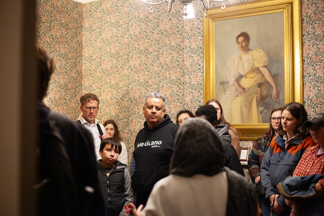 A group of people gathers in a room with patterned wallpaper and a framed portrait of a woman in a yellow dress hanging on the wall; one man wears a black Mexicano hoodie.