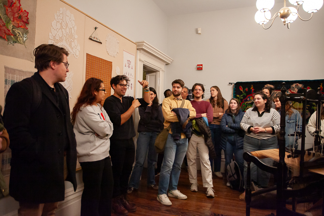 A group of people gathers in a well-lit room, some listening intently while one person in the center gestures as they speak. Various artworks and textiles hang on the walls.