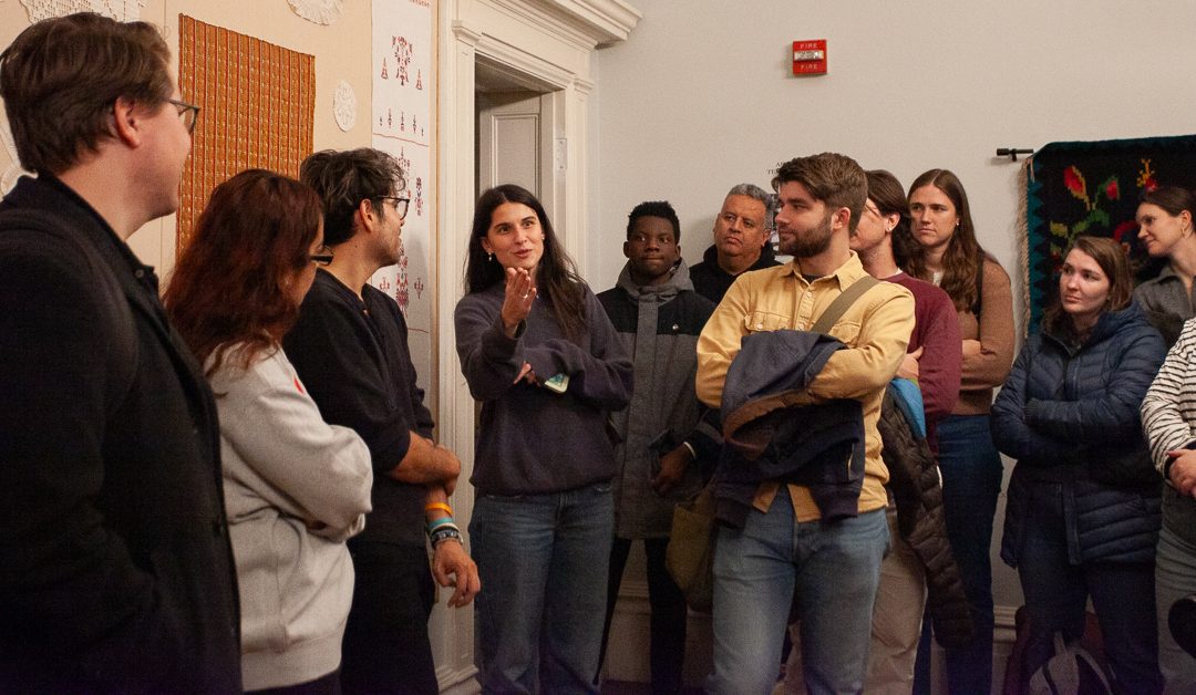 A group of people stand in a room, attentively listening to a woman who is speaking. Some look engaged, while others are thoughtful. Artworks, including textiles, hang on the walls around them.