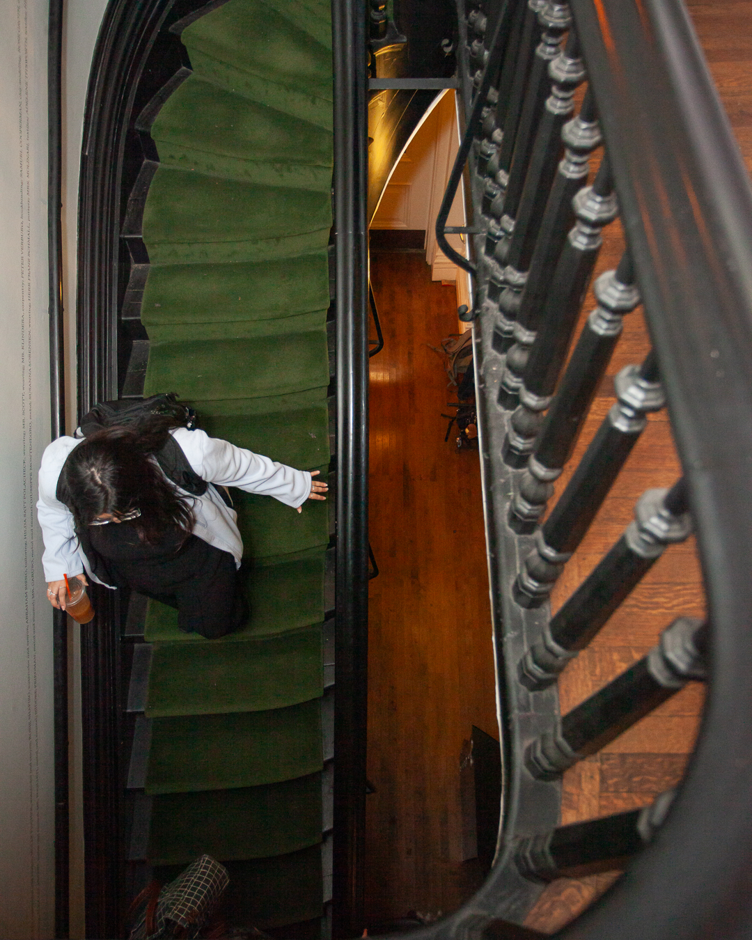 A person with long dark hair, dressed in a white top and black pants, walks up a green-carpeted staircase, seen from above with a curved black railing and a polished wooden floor below.