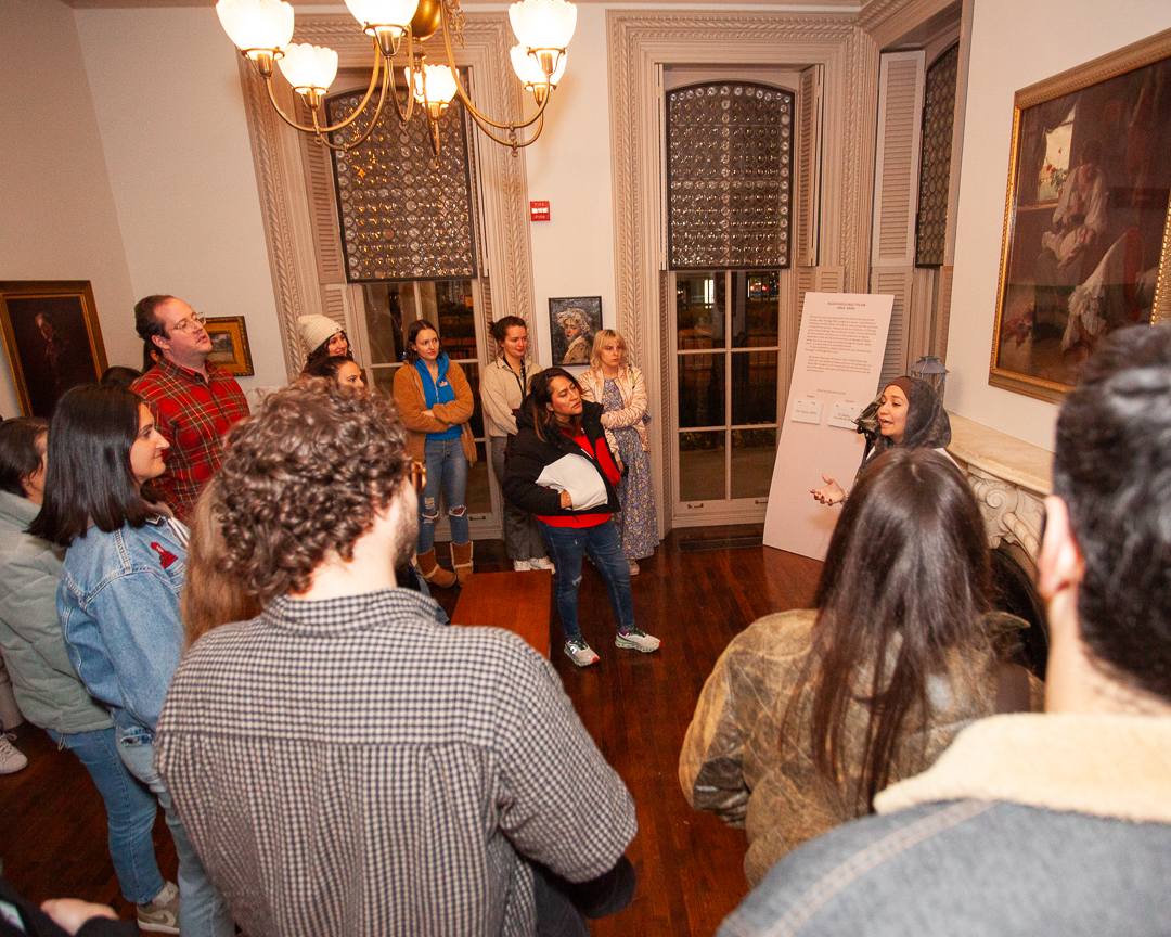 A group of people stands in a well-lit room, listening to a woman speaking near a fireplace. The room features framed artwork, tall windows, wooden floors, and a chandelier hanging from the ceiling.