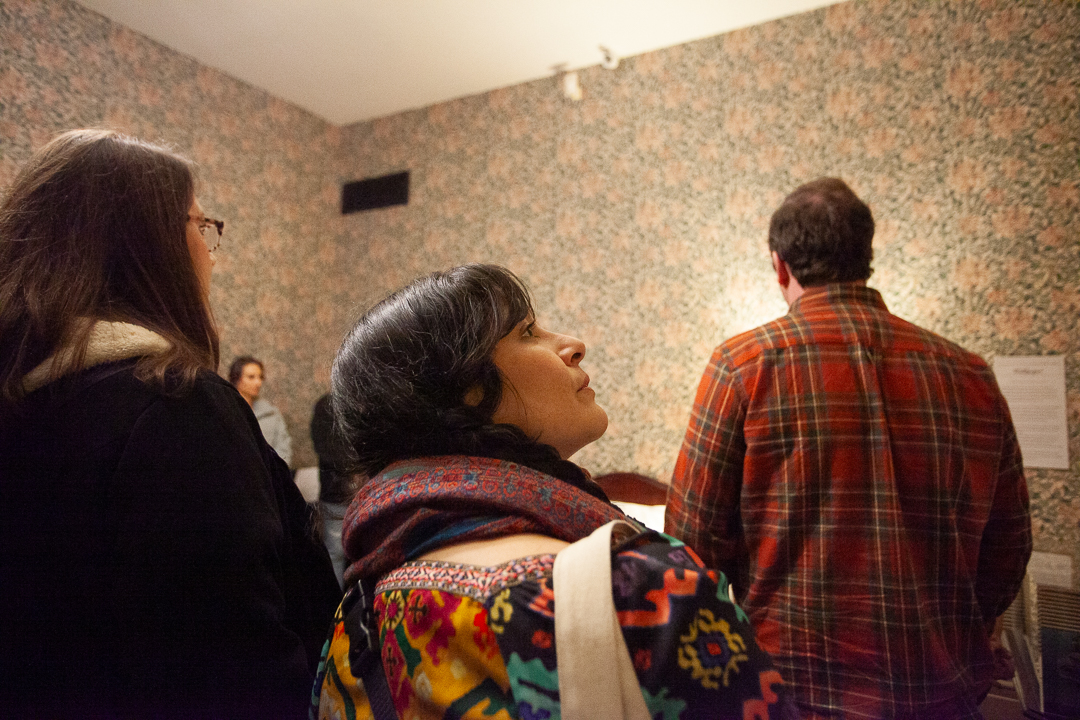 A group of people stand in a dimly lit room with floral wallpaper, some facing away from the camera while one woman in the center looks upward, wearing a colorful scarf and patterned top.