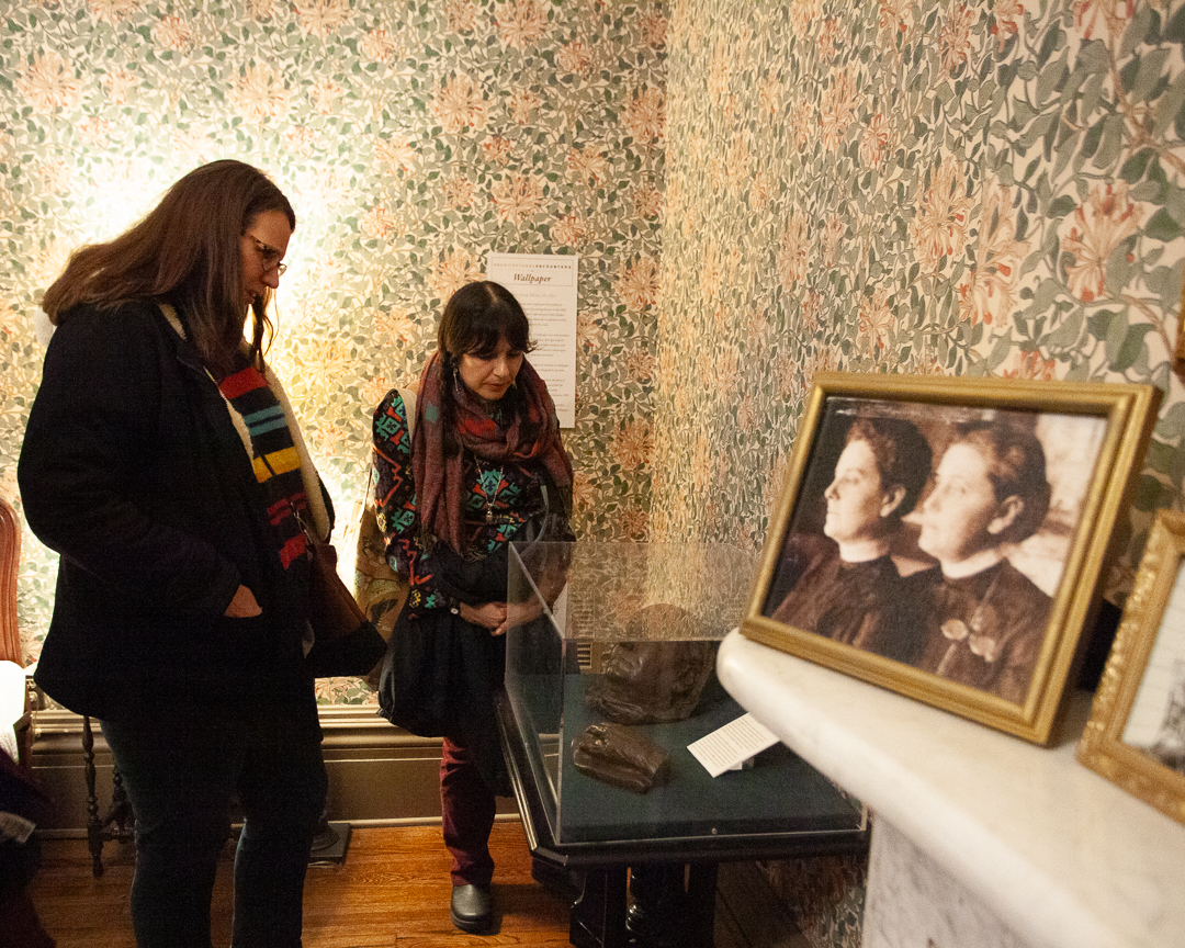 Two women stand in a museum room with floral wallpaper, looking at historical artifacts and a framed old photograph of two women on a table. One artifact is displayed in a clear case.