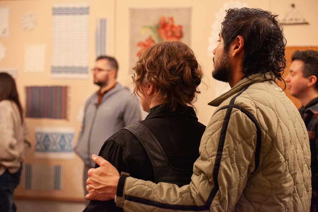 A man and woman stand close together, with his arm around her shoulders, looking at artwork on a wall. Other people are also observing the displayed textile pieces in a warmly lit gallery space.