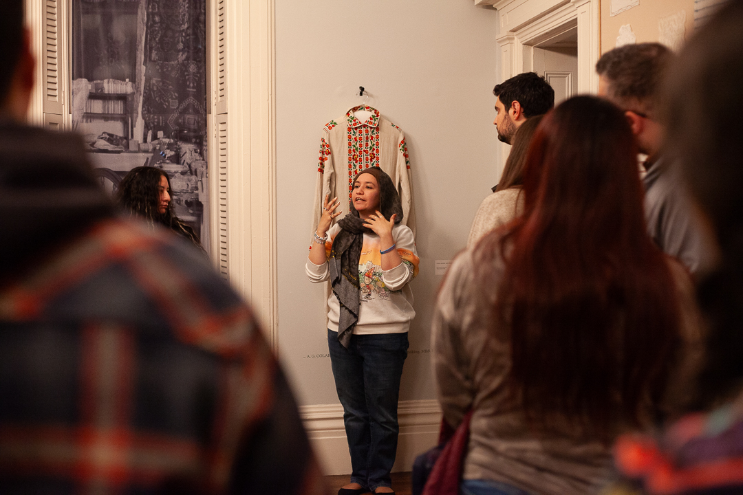 A woman stands in front of a group, gesturing as she speaks in a room with ornate details. Behind her, an embroidered garment is displayed on the wall. The group listens attentively.