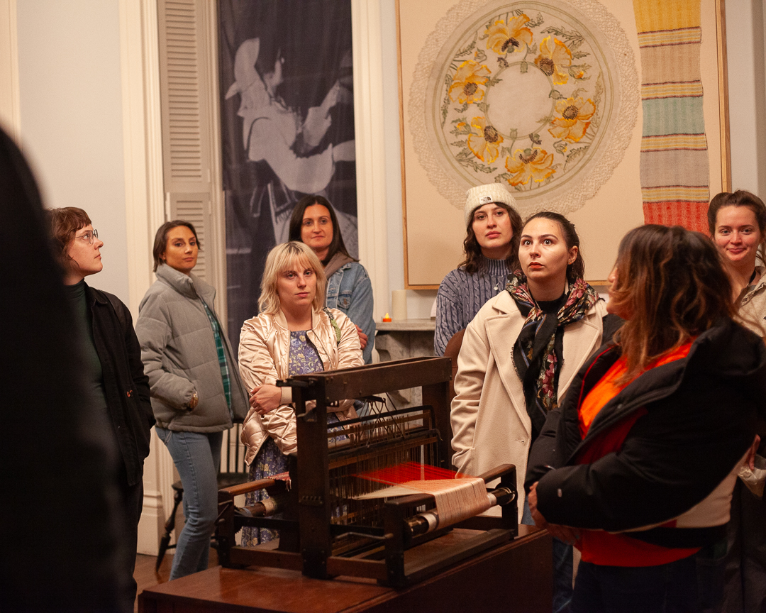 A group of women stand around a wooden weaving loom in a museum gallery, observing attentively. Textile art and a large fabric with a floral design hang on the walls behind them.
